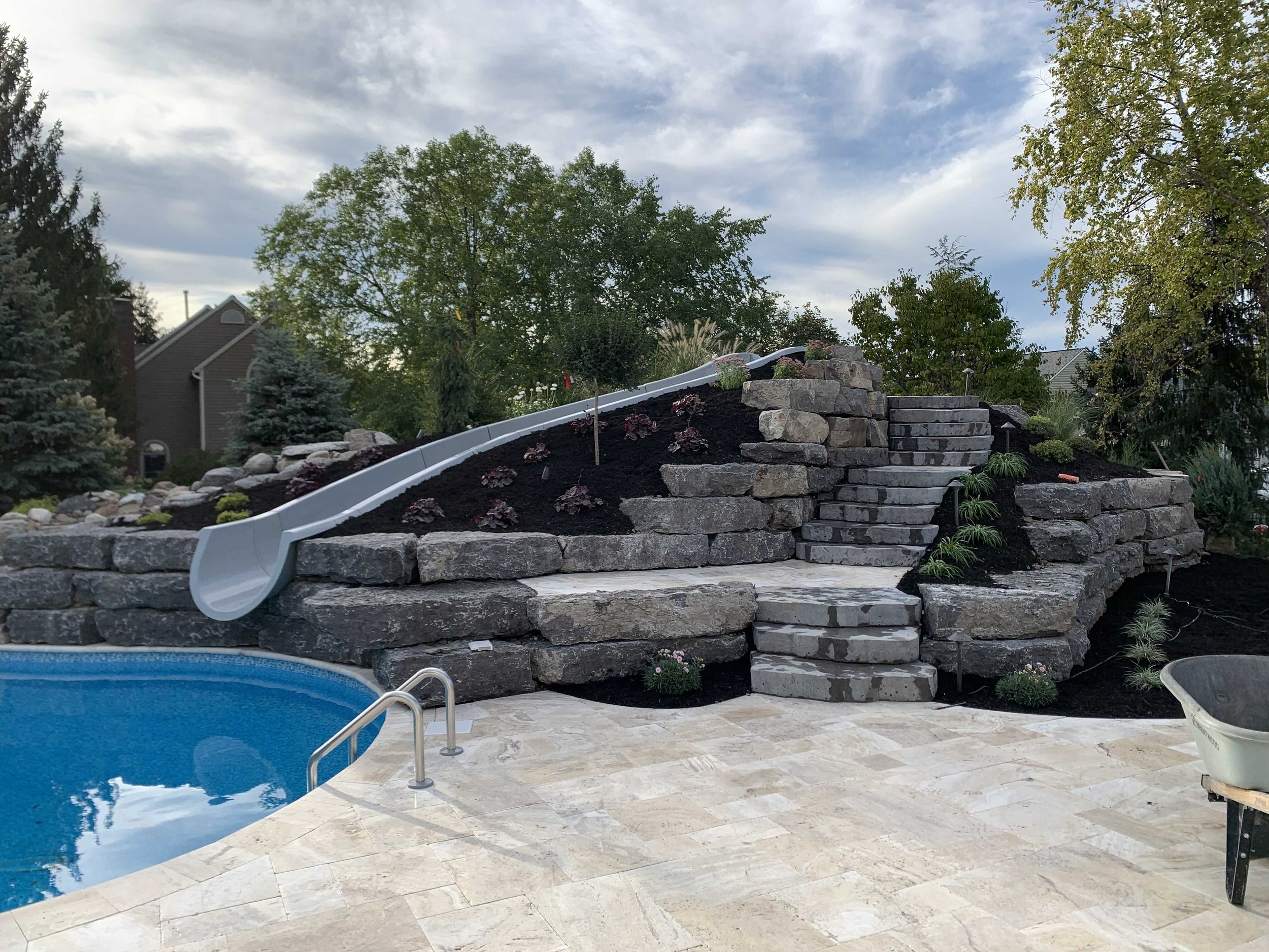A backyard scene with a swimming pool, a stone retaining wall, a water slide, and landscaped plants and trees.