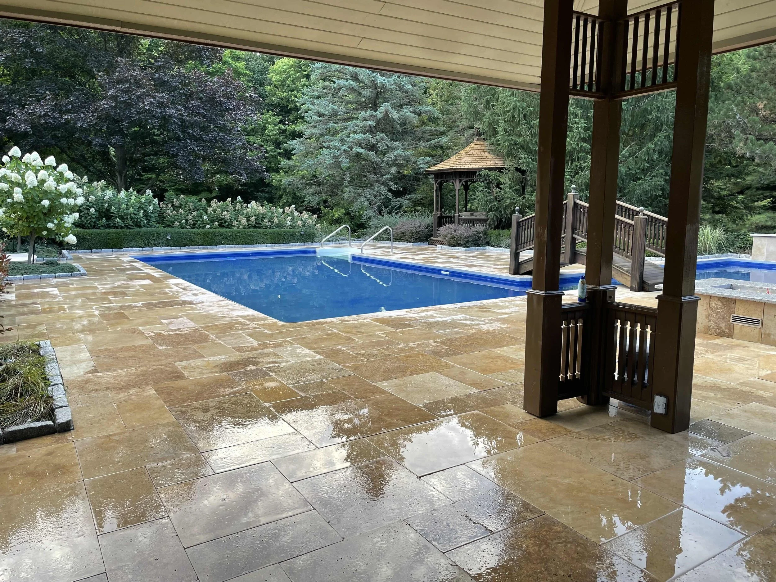 Empty backyard swimming pool with a stone deck, surrounded by lush trees and bushes, and a small wooden gazebo in the background.