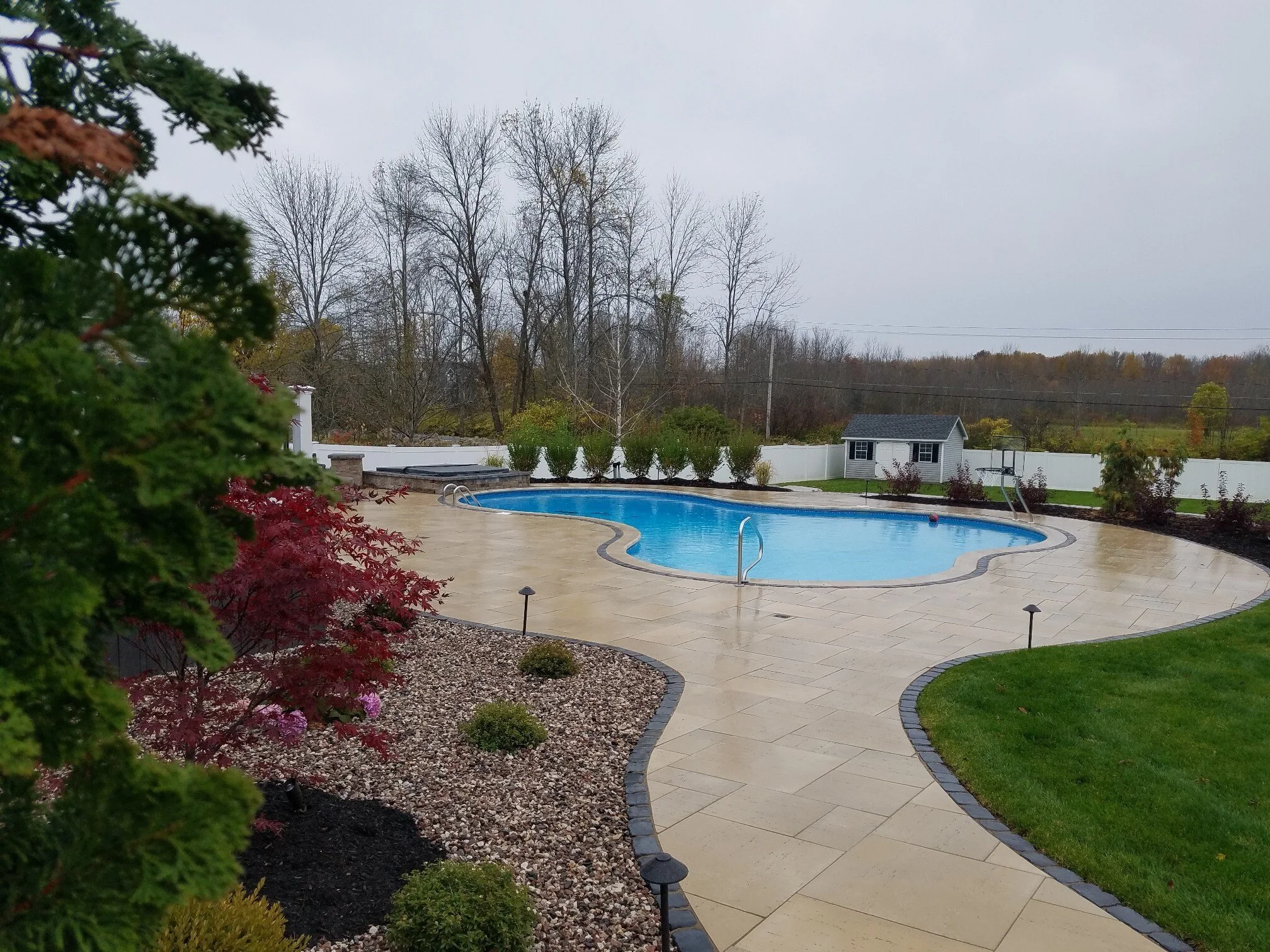 An outdoor pool surrounded by a beige paved walkway, with a small shed in the background and deciduous trees beyond a white fence on an overcast day.