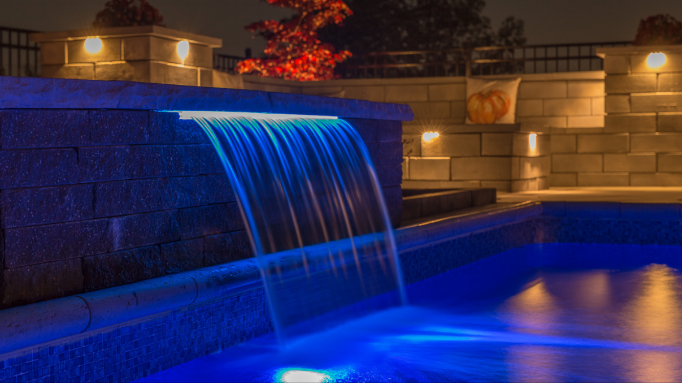 Night view of a backyard with a lit water feature illuminated in blue, brick walls with warm lighting, and a cushion with an orange pumpkin design in the background.