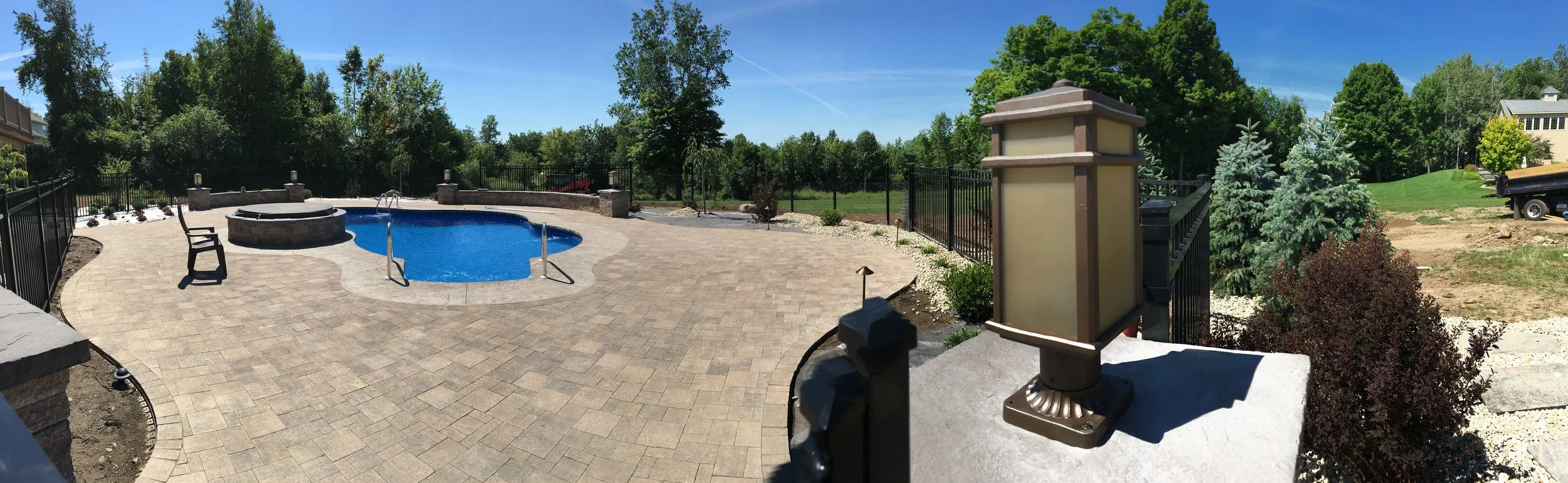 Residential backyard with a fenced-in swimming pool, brick paver patio, a fire pit, surrounded by trees and shrubs, and a house under construction with dirt and grass area nearby.