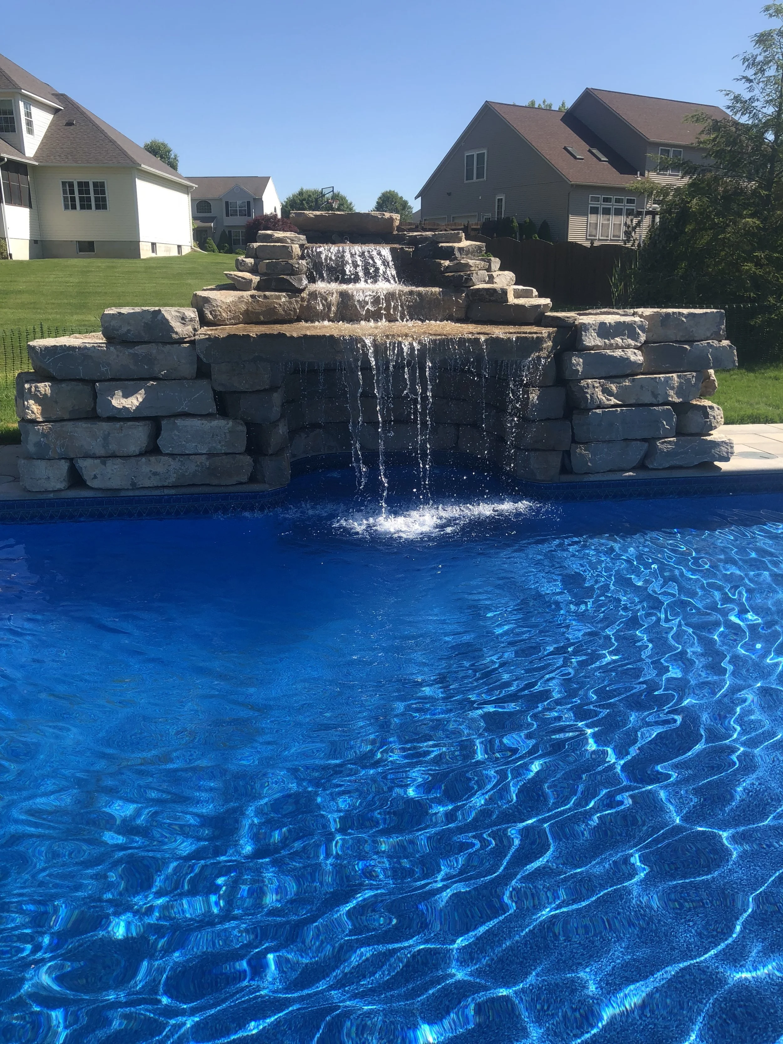 A backyard with a stone waterfall feature flowing into a blue swimming pool, with houses and green lawns in the background under a clear sky.