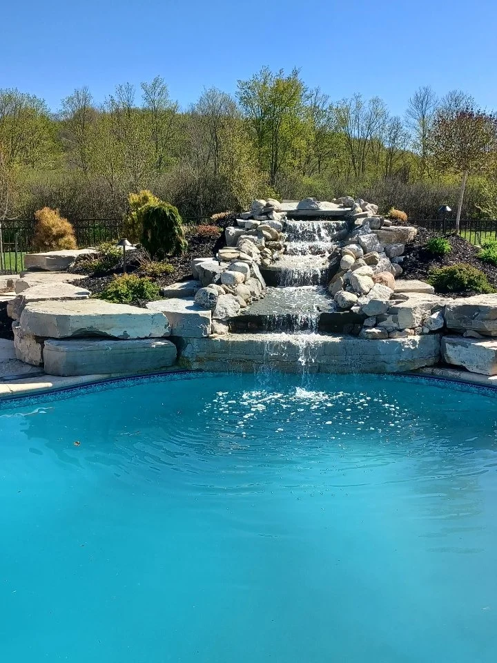 A swimming pool with a waterfall feature made of rocks, surrounded by greenery and trees under a clear blue sky.