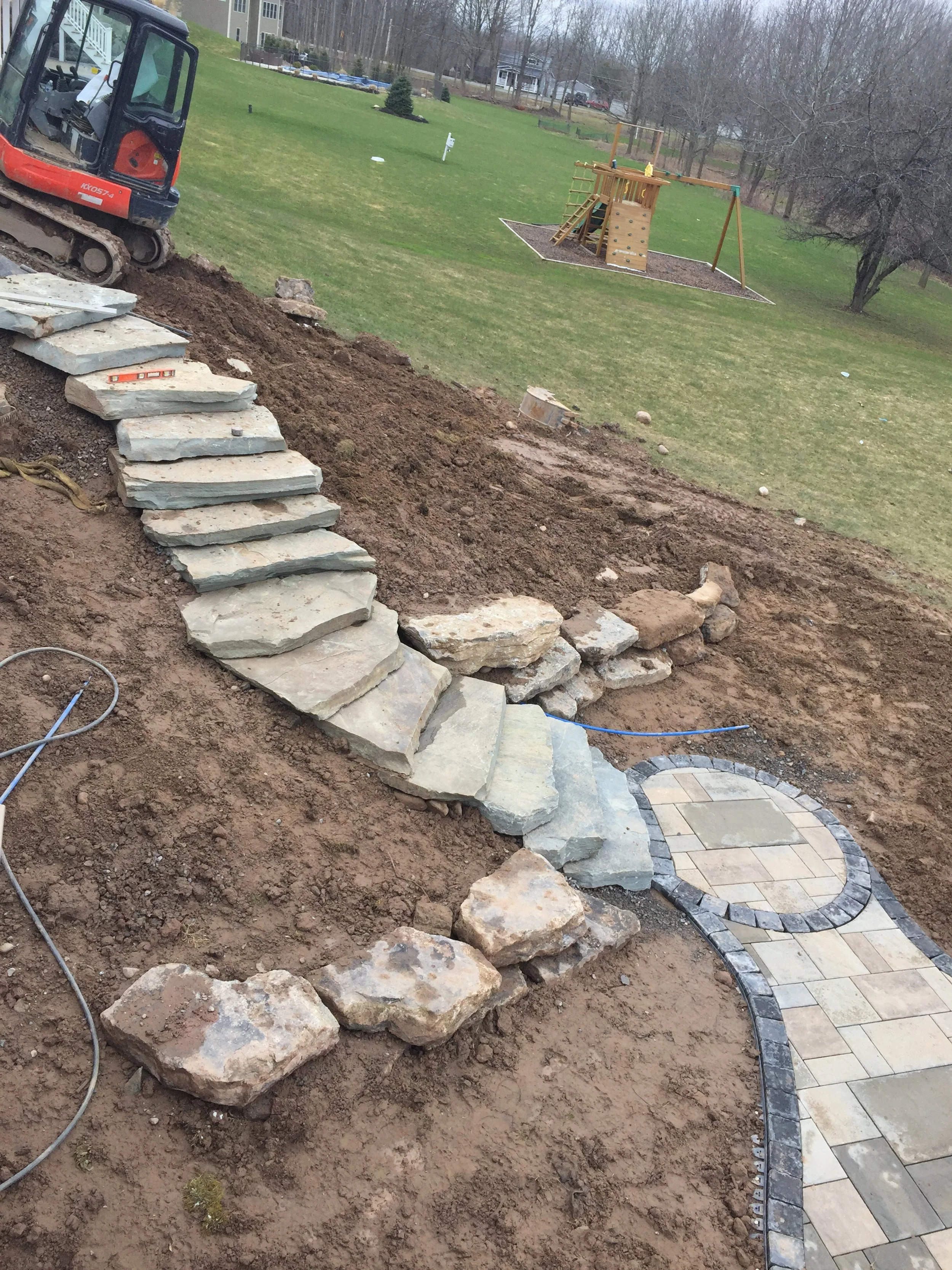 Stone stairs leading up a slope next to a winding paved pathway in a backyard, with a small construction vehicle on the left and a wooden playset with swings and a climbing wall in the background.