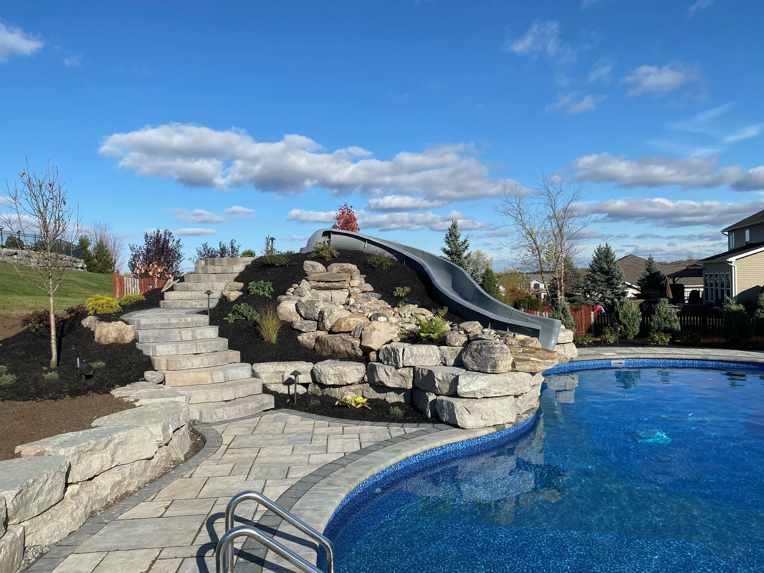 Residential backyard with a swimming pool, stone slide, and landscaped garden under a partly cloudy sky.