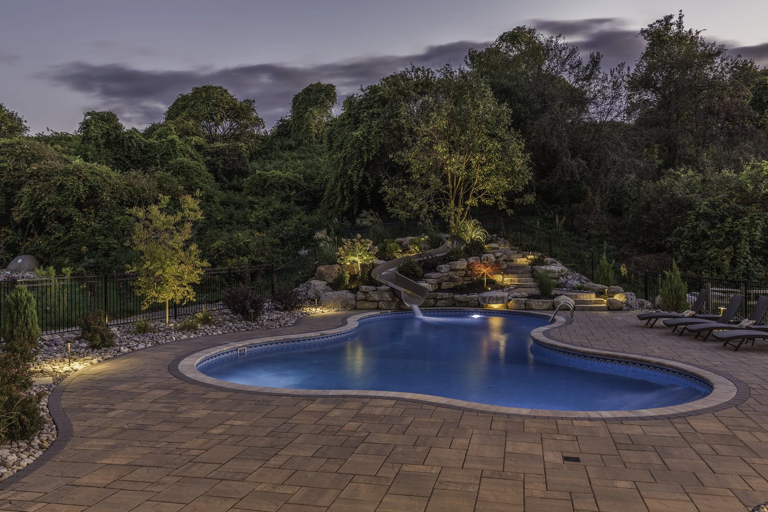A backyard swimming pool area at dusk with a slide, lounge chairs, and landscaped garden with trees and rocks.