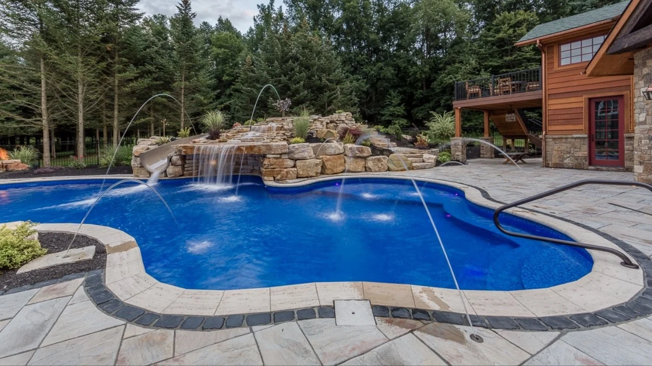 A backyard swimming pool with a stone waterfall feature, surrounded by a stone patio, with a wooden house and a wooded area in the background.