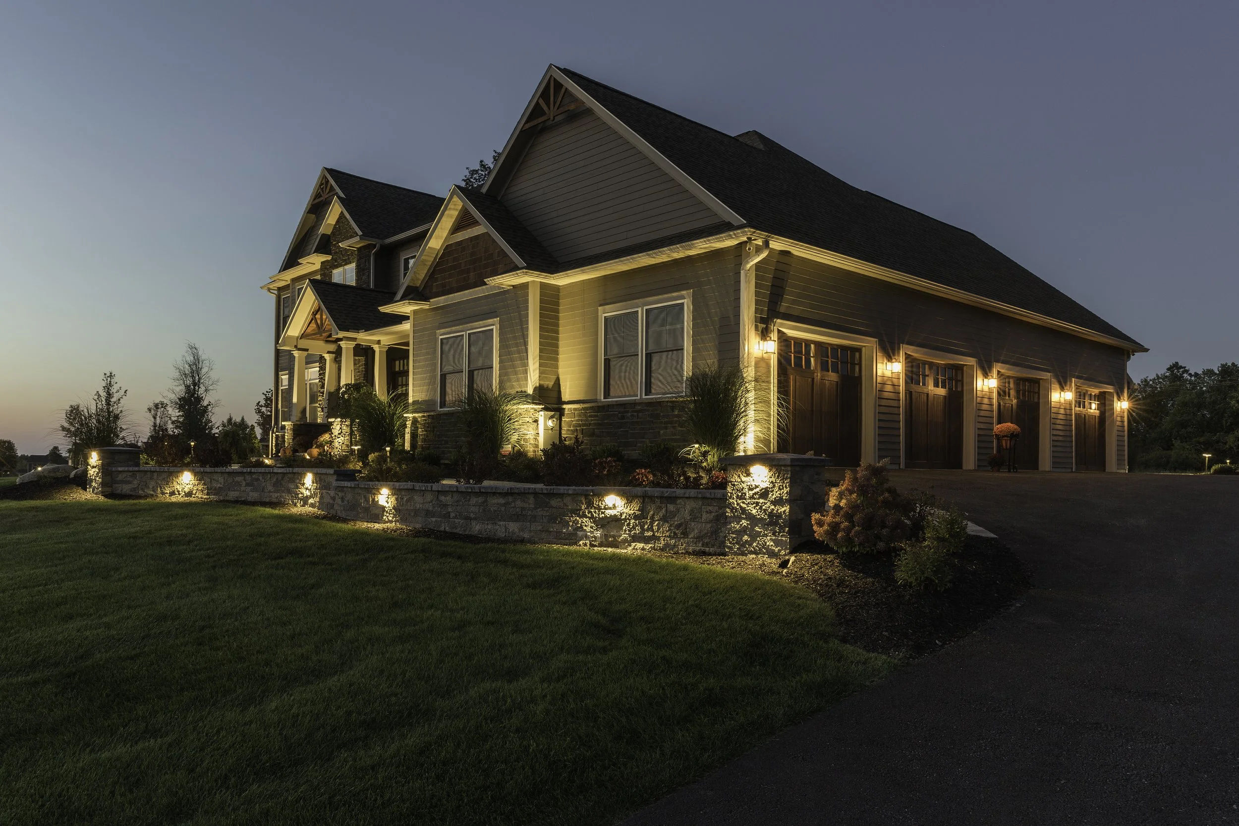 A large residential house at dusk illuminated by exterior lighting, with a well-manicured lawn, a stone retaining wall, and a paved driveway.