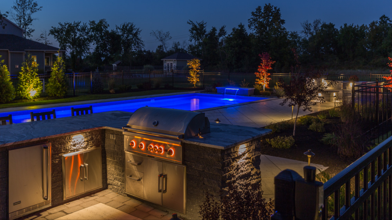Nighttime view of a backyard with a lit swimming pool, outdoor kitchen with grill, and landscaped garden with trees and bushes.