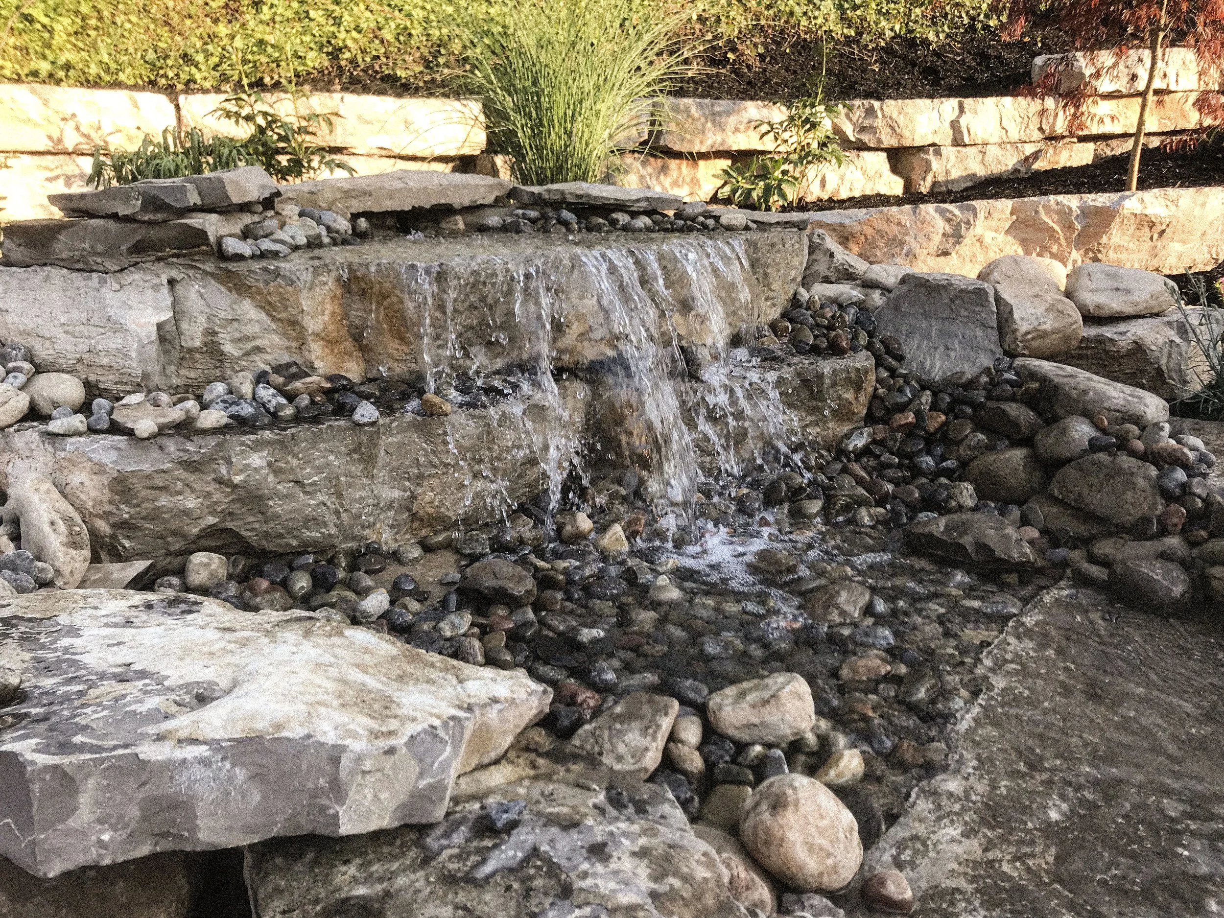 Small waterfall cascading over stacked stones in a landscaped garden with decorative rocks and plants.
