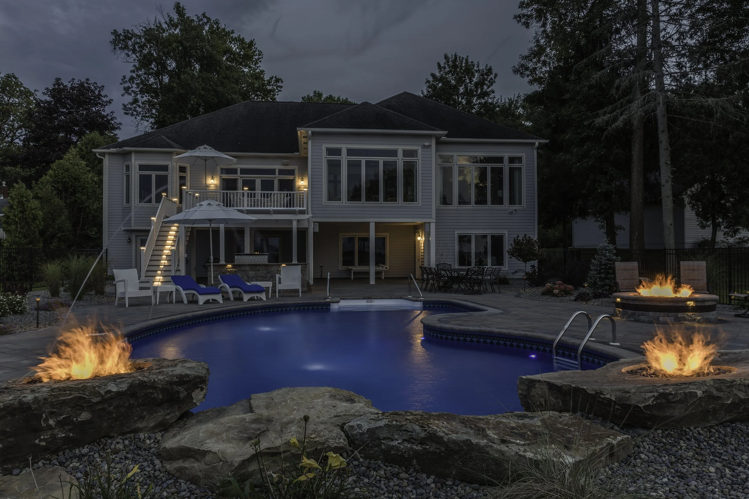 Nighttime view of a backyard swimming pool with two fire features on rocks, outdoor seating, and a large house with multiple windows and balconies in the background.