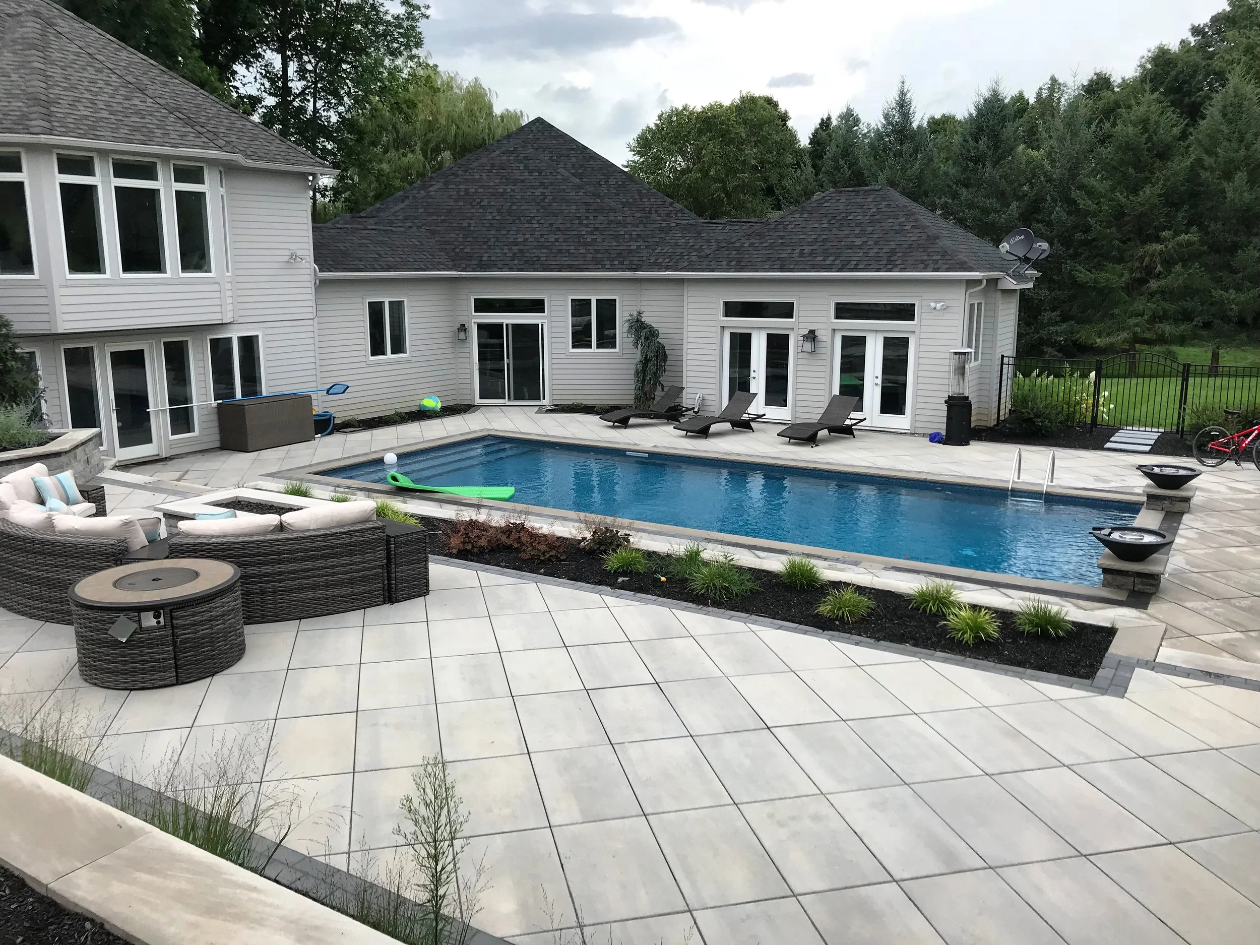 Backyard with a swimming pool surrounded by patio furniture and a house with multiple windows and doors, some with loungers and outdoor decor, and surrounded by trees.