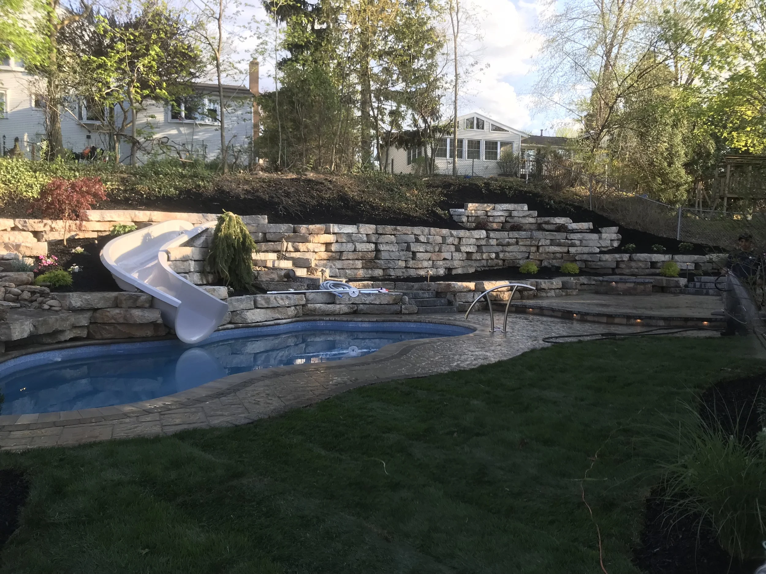 Residential backyard with a swimming pool featuring a white slide, stone retaining wall, and landscaping with trees and bushes.