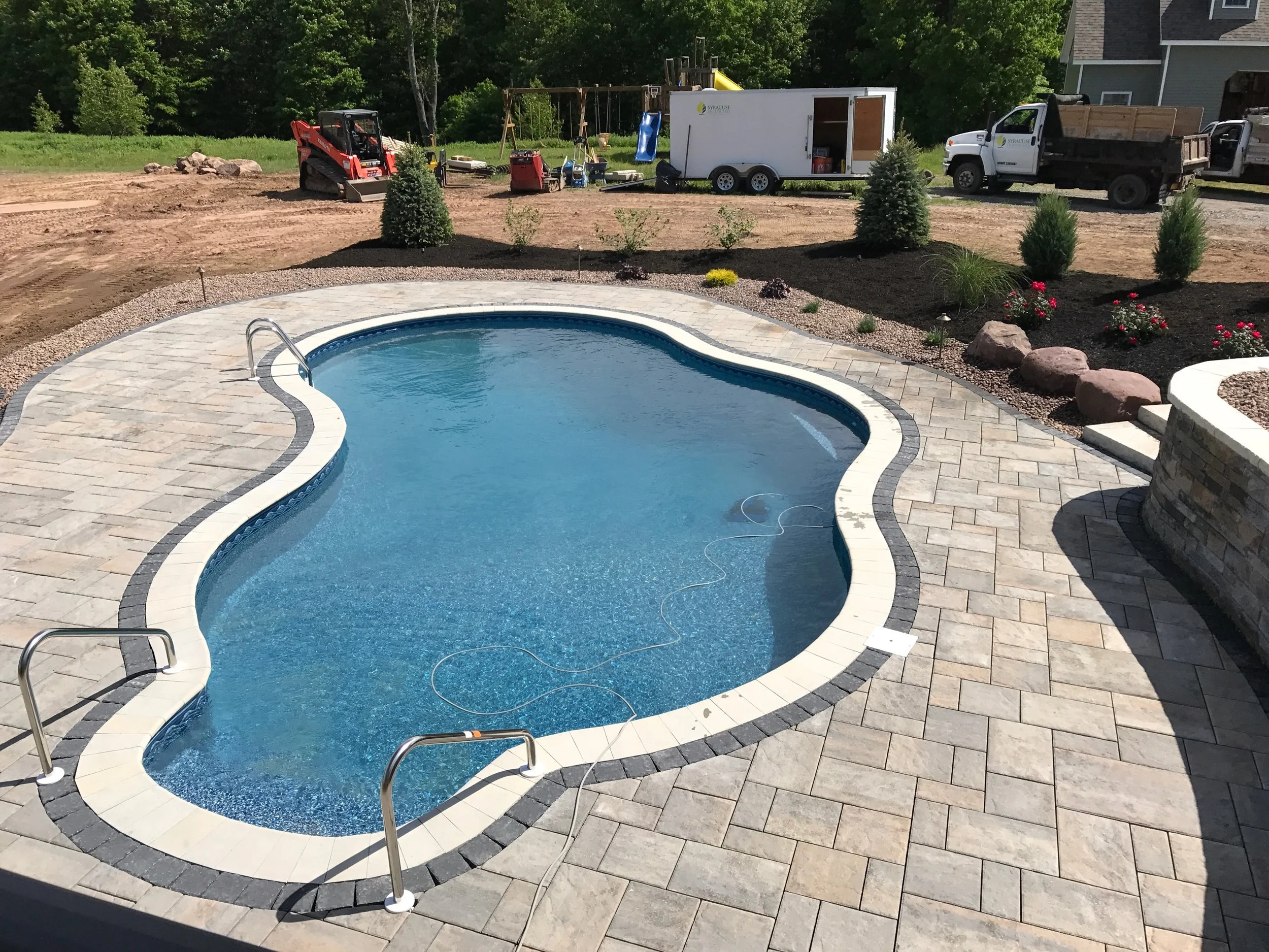 Empty swimming pool with stone paving, newly landscaped backyard, construction tools, gravel, trucks, and trailer in background.