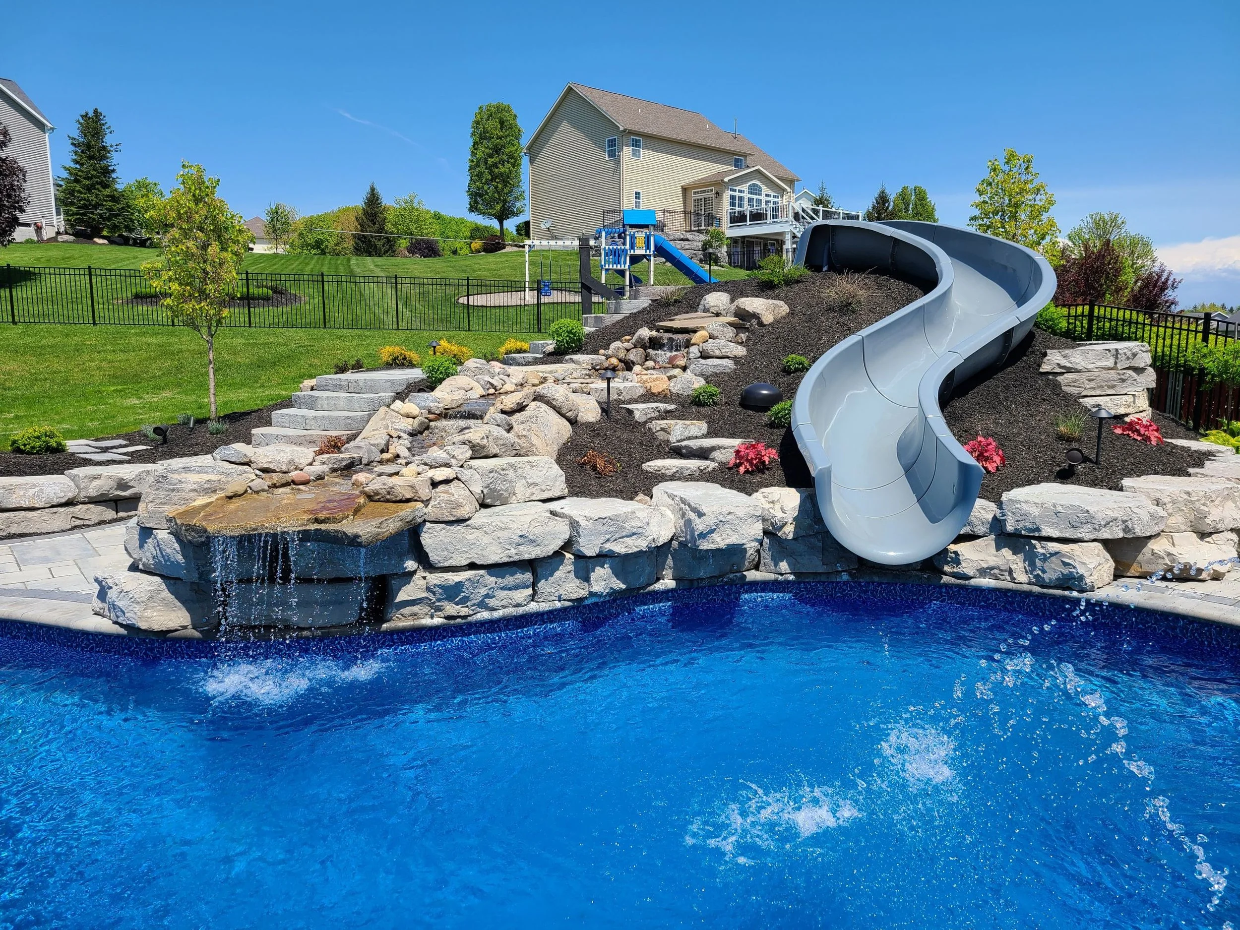 A backyard with a swimming pool, a rock waterfall feature, and a waterslide, surrounded by a stone patio and a well-maintained lawn with trees and shrubs.