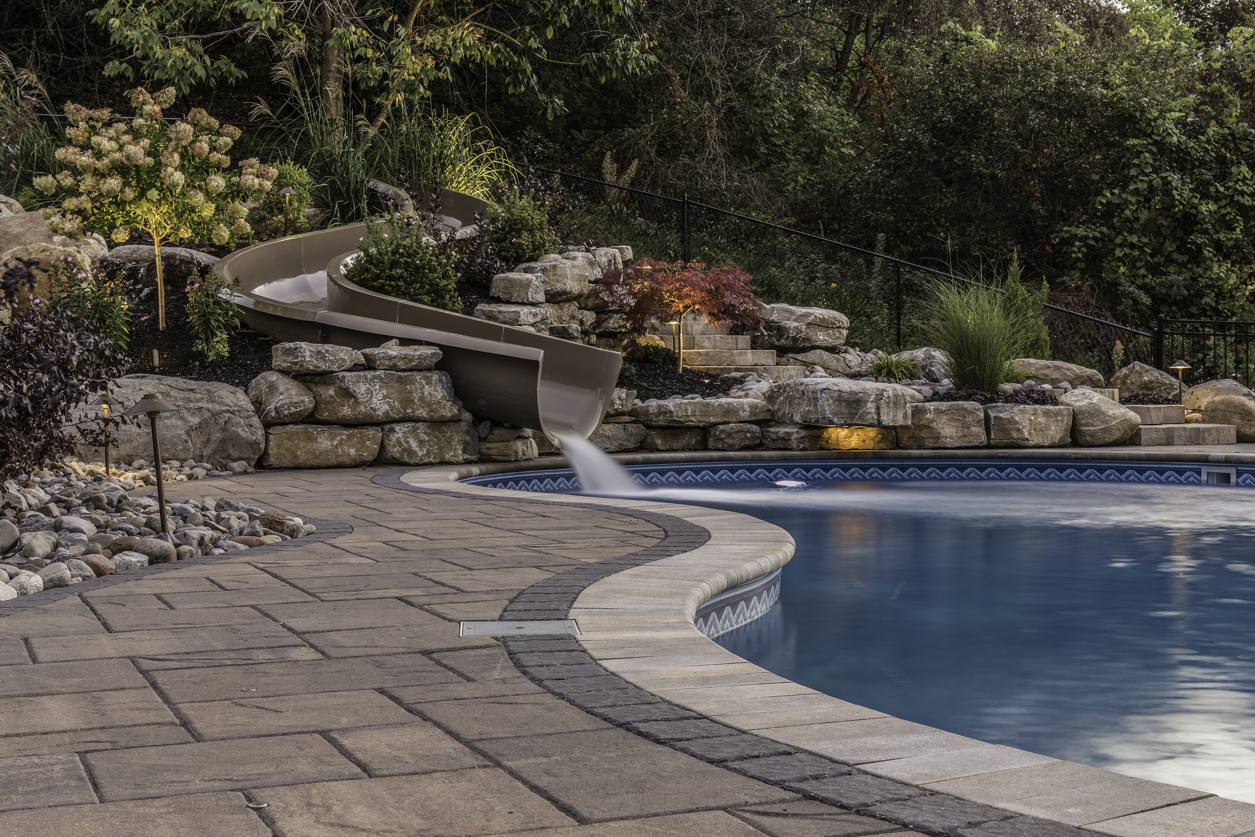 A backyard swimming pool with a water slide, surrounded by stone and brick patio, and landscaped with trees, shrubs, and decorative rocks.
