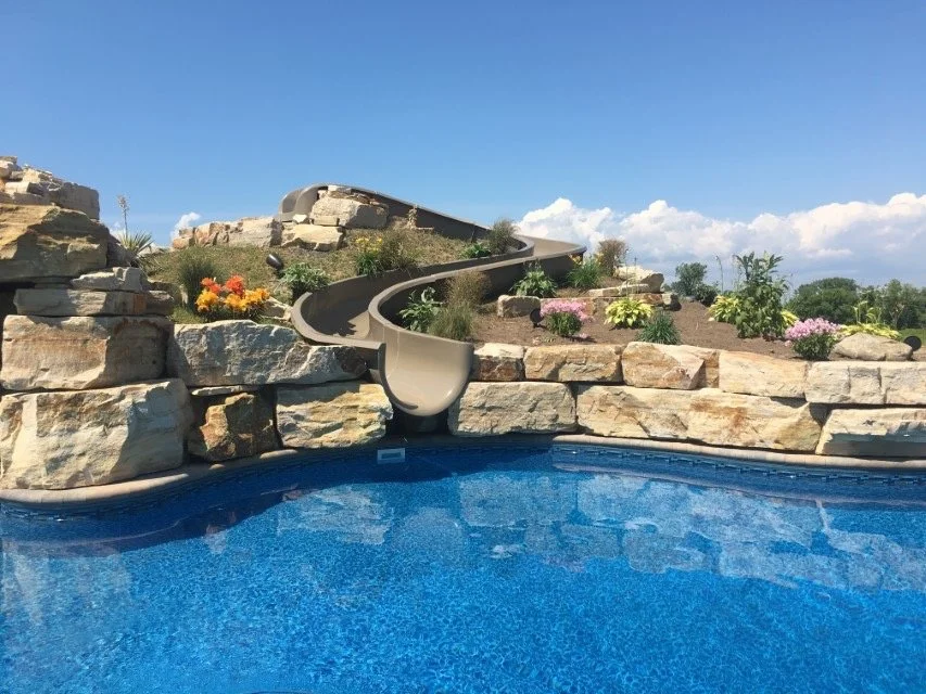 A swimming pool with a rock waterfall and a waterslide against a backdrop of a blue sky with some clouds.