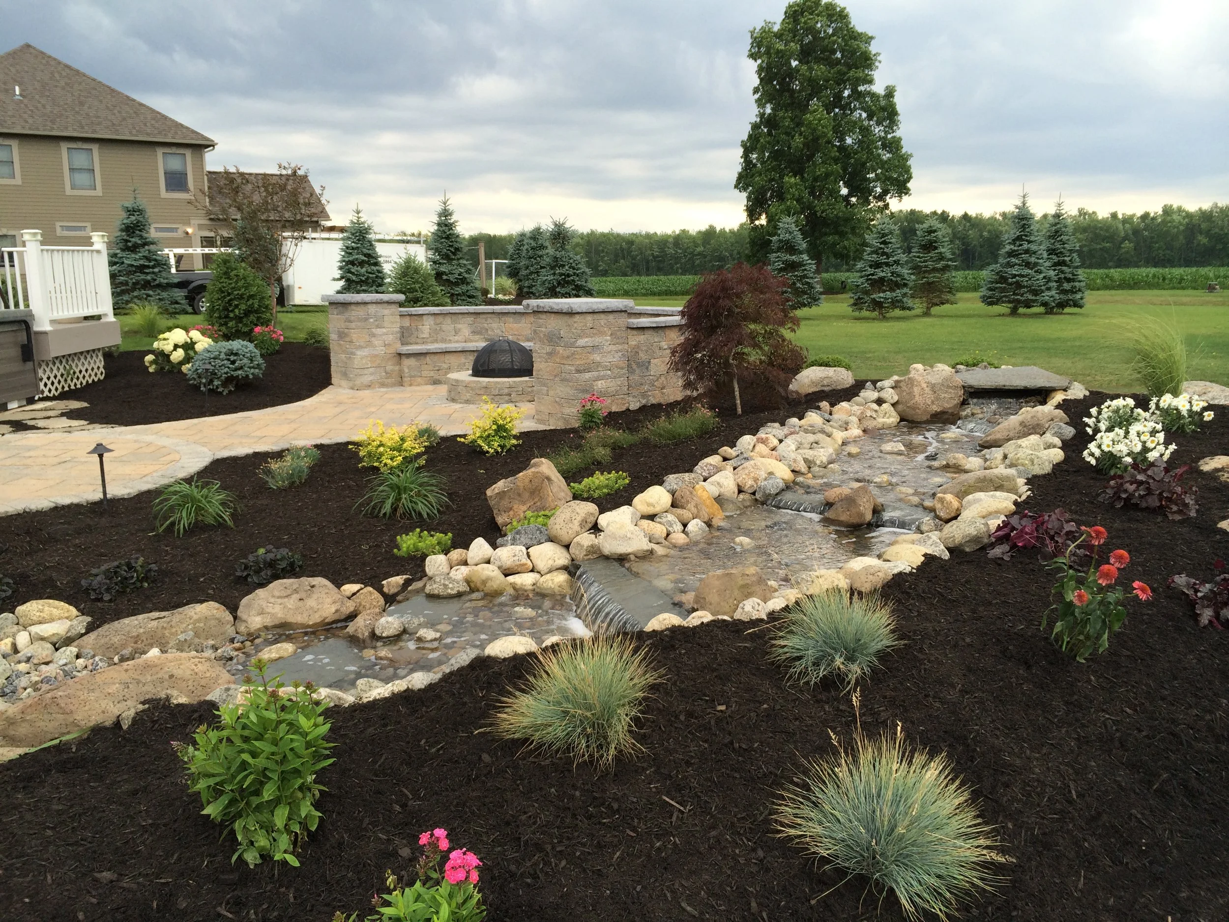 A landscaped backyard with a stone patio, a small waterfall, and a dry creek bed. There are various plants, flowers, and small shrubs, with a large tree in the background and a row of evergreen trees beyond the lawn.
