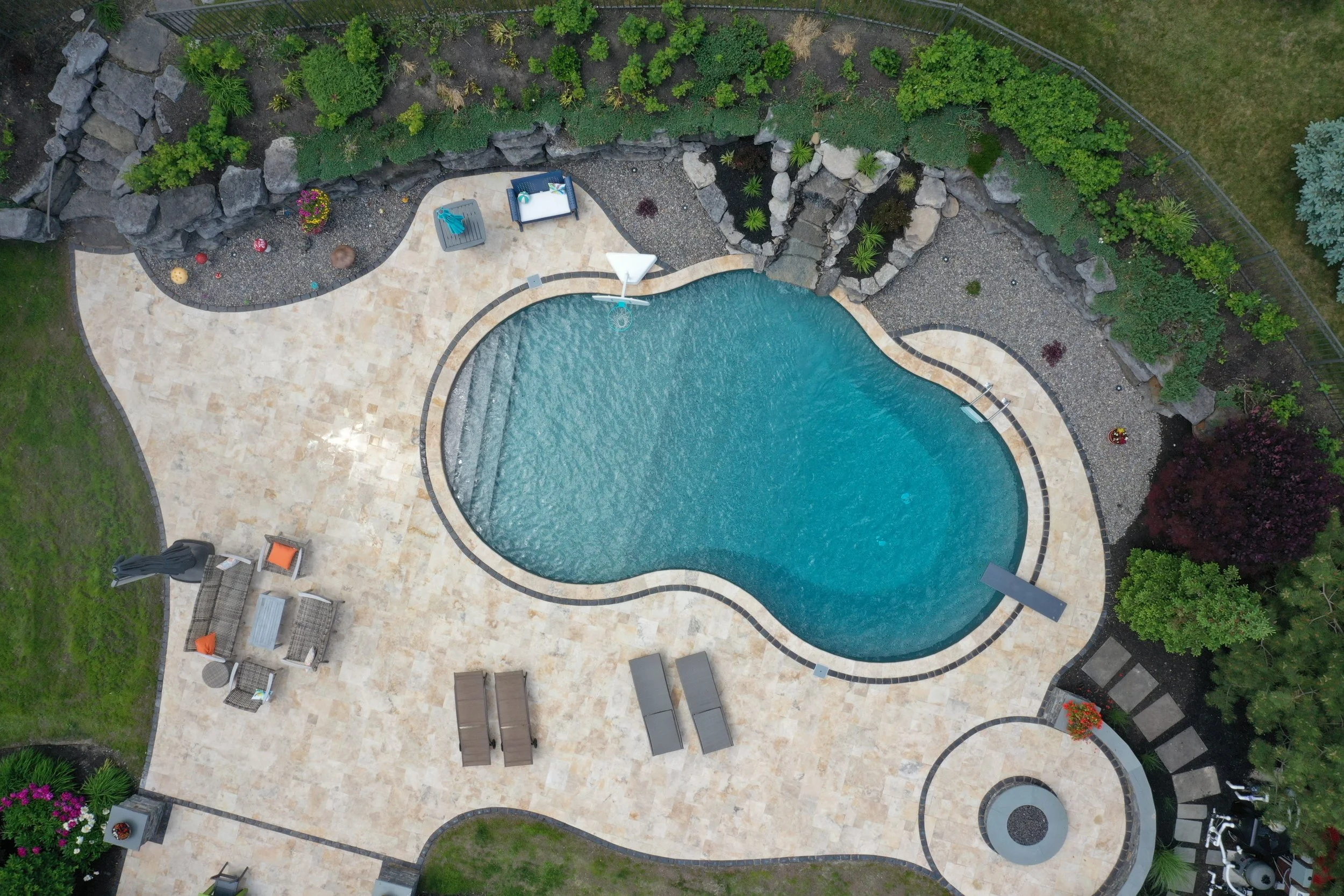 A backyard with a kidney-shaped swimming pool surrounded by a beige tiled deck. There are outdoor furniture pieces including a seating area with four chairs and a small table, two lounge chairs, and a small sitting area with two cushions. The yard is bordered by landscaped garden beds with various plants, rocks, and trees, and there is a grassy area to the left.