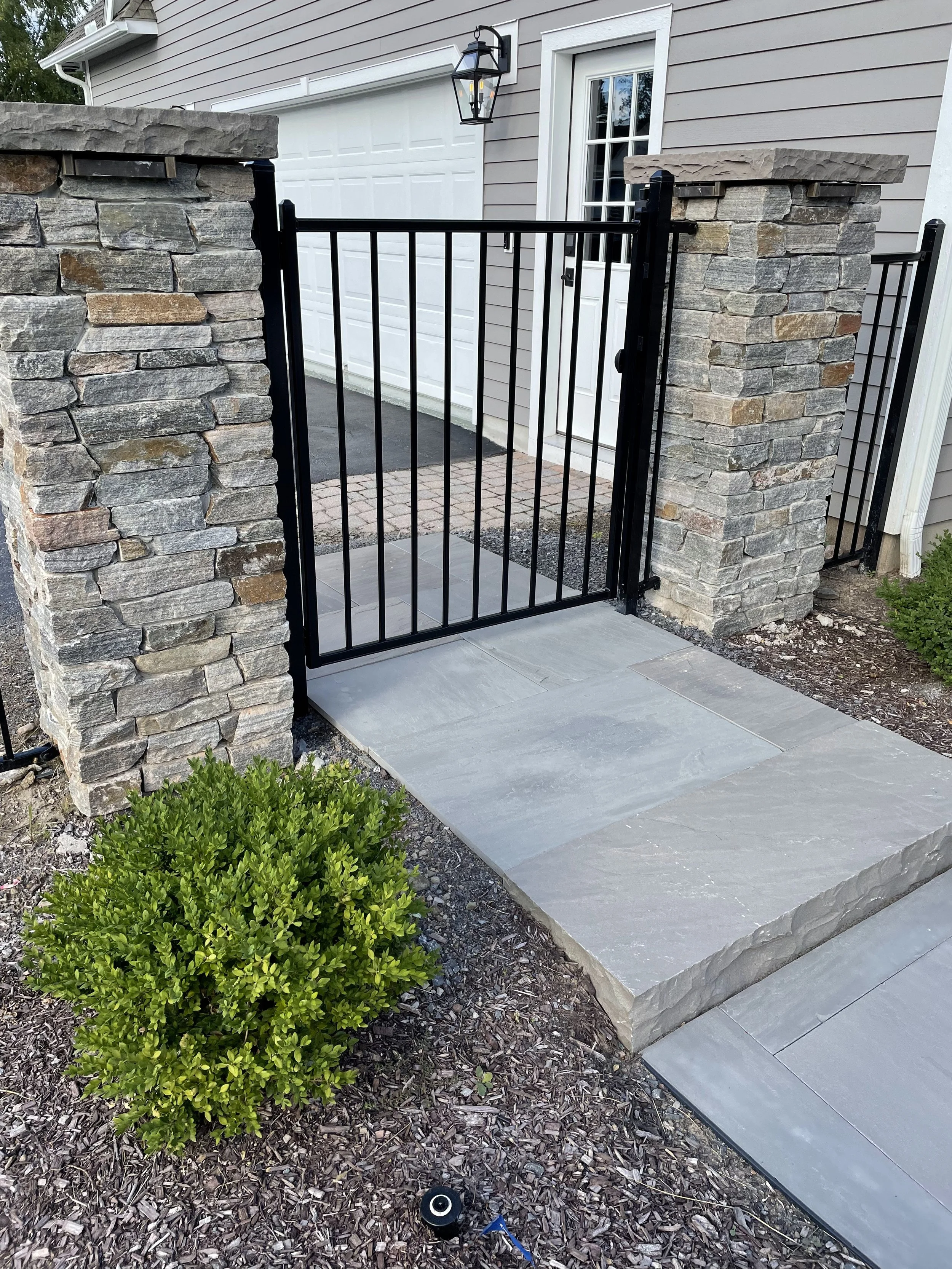 A black metal gate with vertical bars, flanked by two stone pillars, leading to a driveway and outdoor area next to a house with light gray siding, a white door, and a mounted black lantern-style light fixture.