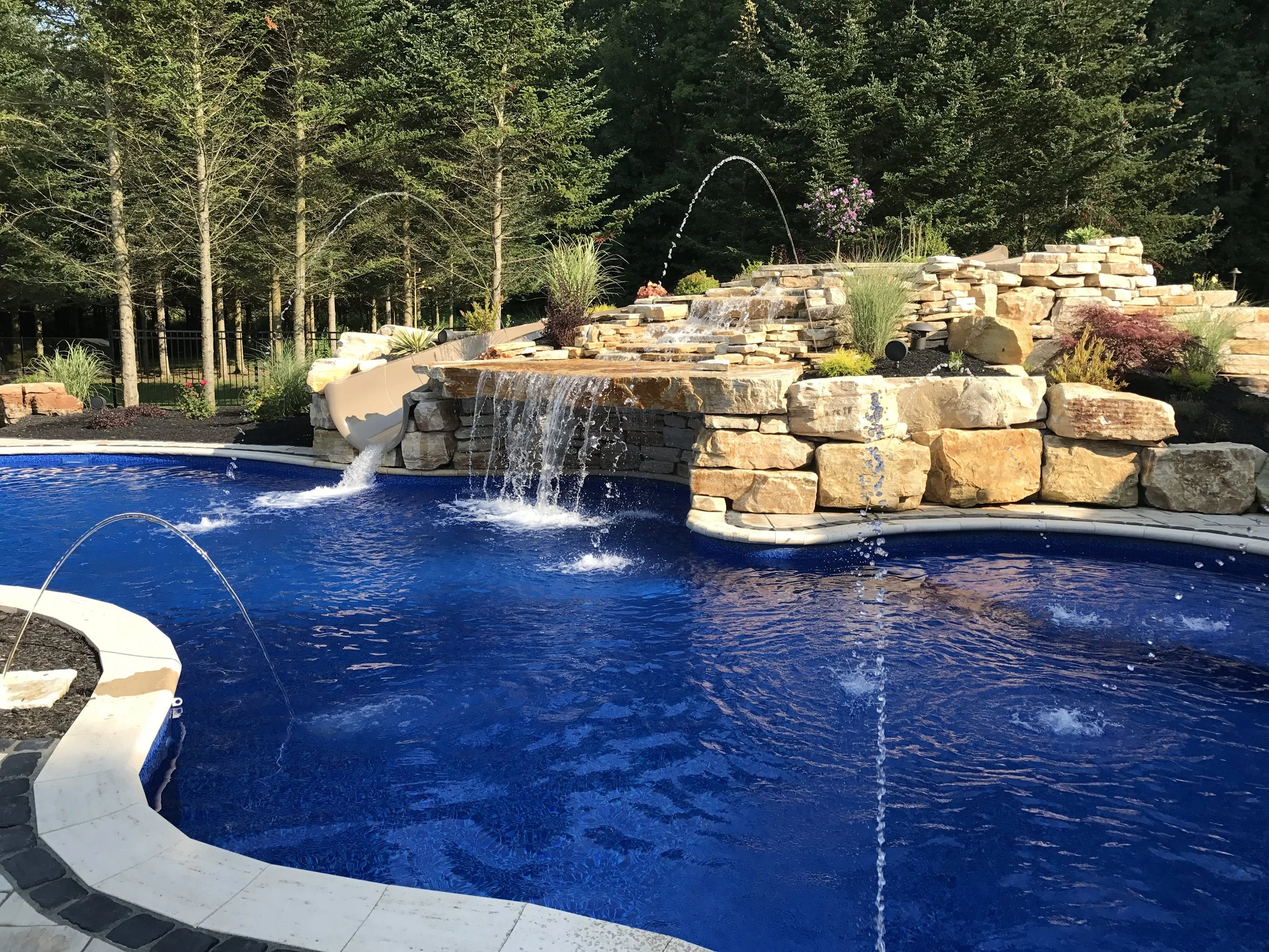 Backyard swimming pool with a rock waterfall feature and lush greenery in the background.