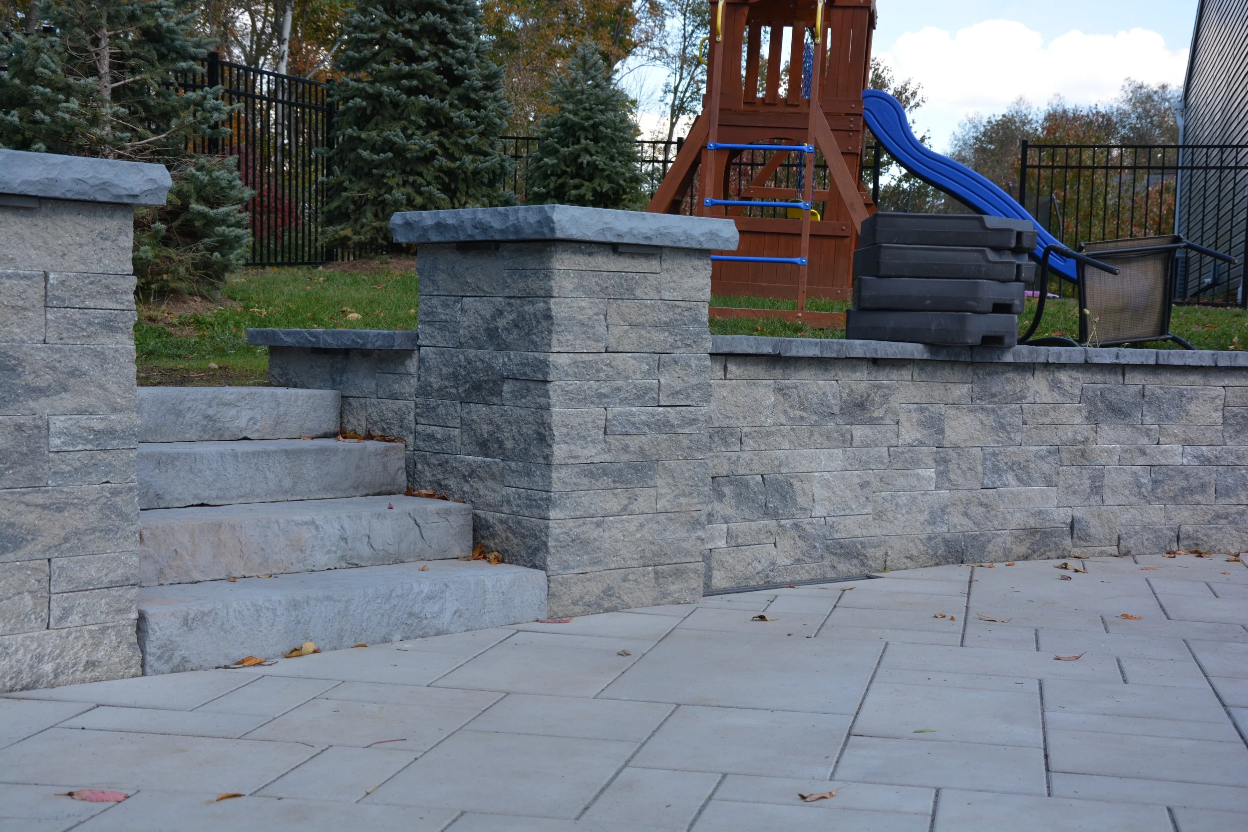 Outdoor backyard patio with stone steps, stone wall, and a playground with a blue slide, black stacking blocks, and a wooden play structure, surrounded by trees and a black metal fence.