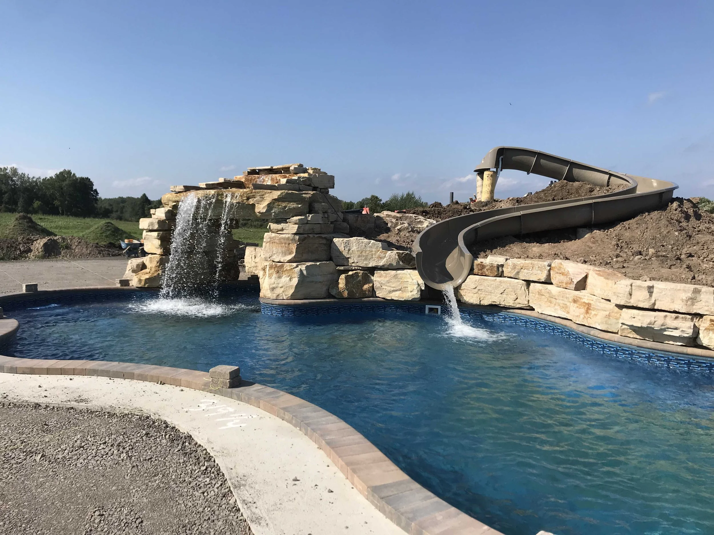 A backyard swimming pool with a waterfall feature and a waterslide under construction, surrounded by dirt and gravel.