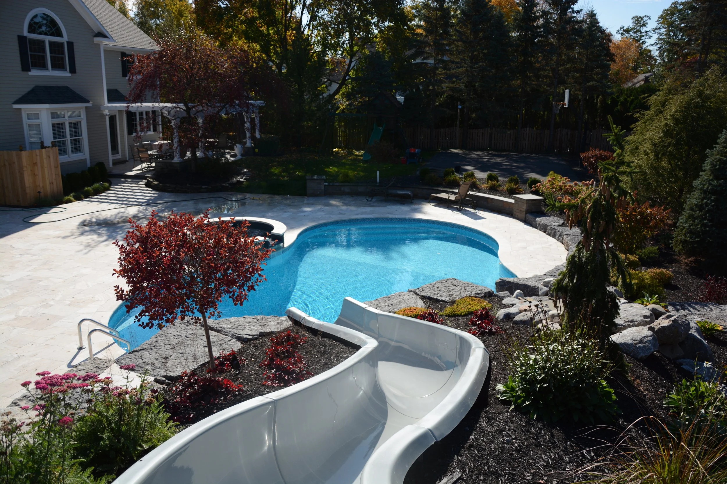 A backyard with a swimming pool, a hot tub, a water slide, and surrounding landscaped greenery and trees.