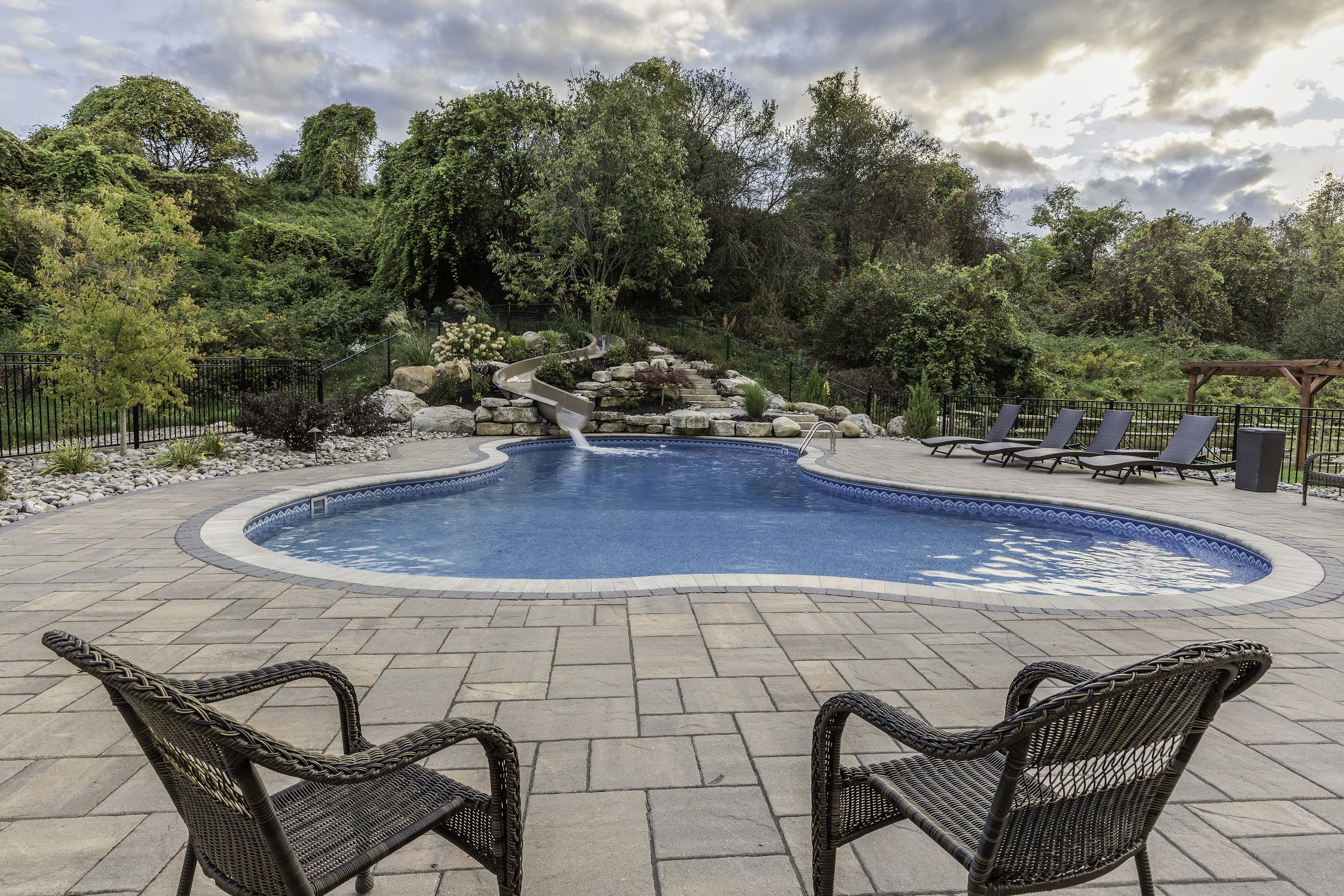 A backyard swimming pool with a slide and waterfall feature, surrounded by lounge chairs and lush greenery, under a partly cloudy sky.