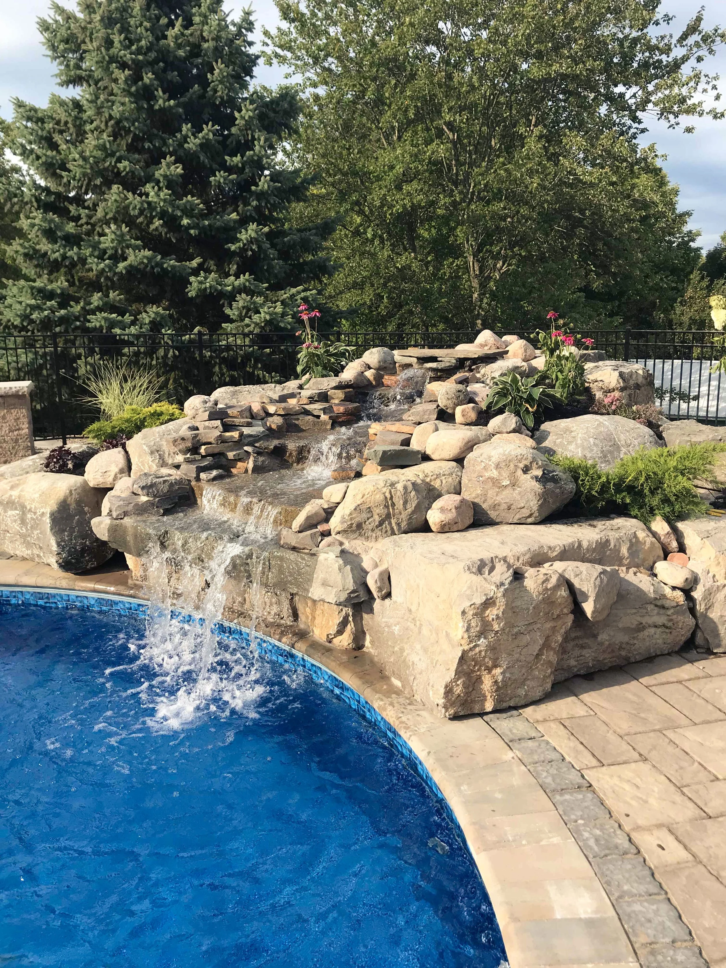 Waterfall feature with rocks and plants next to a swimming pool, with trees and a cloudy sky in the background.