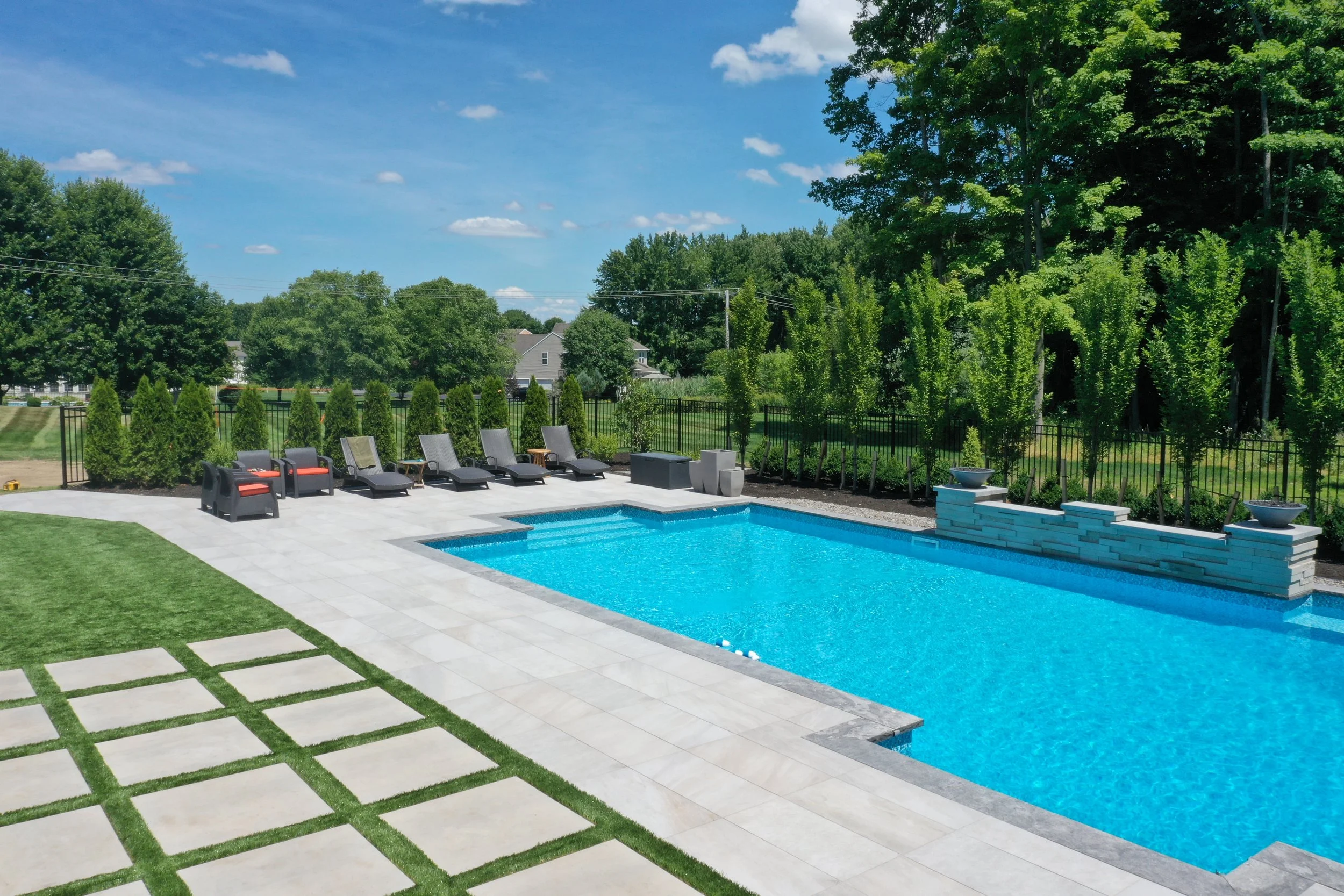 A backyard with a swimming pool, lounge chairs, and a landscaped garden with trees and a black fence under a blue sky with some clouds.