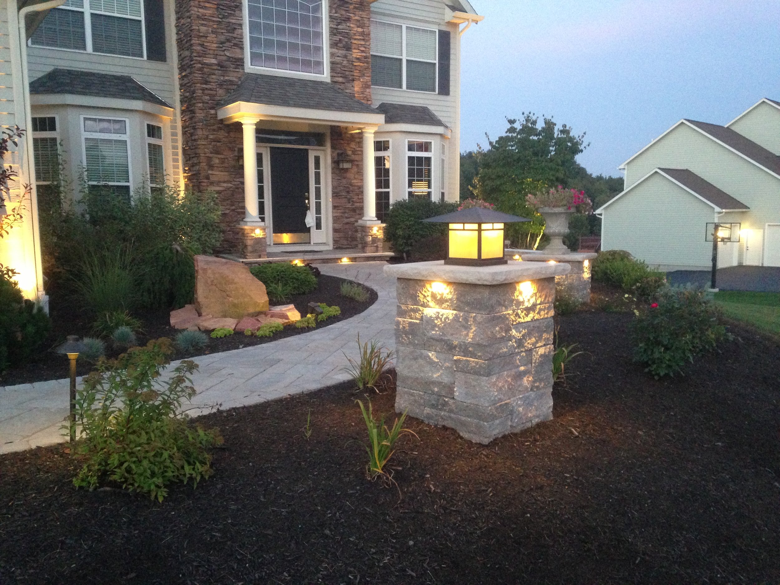 Beautiful front yard of a house illuminated at dusk, featuring a stone pathway, garden beds, outdoor lighting, and a large stone pillar with a lantern on top.