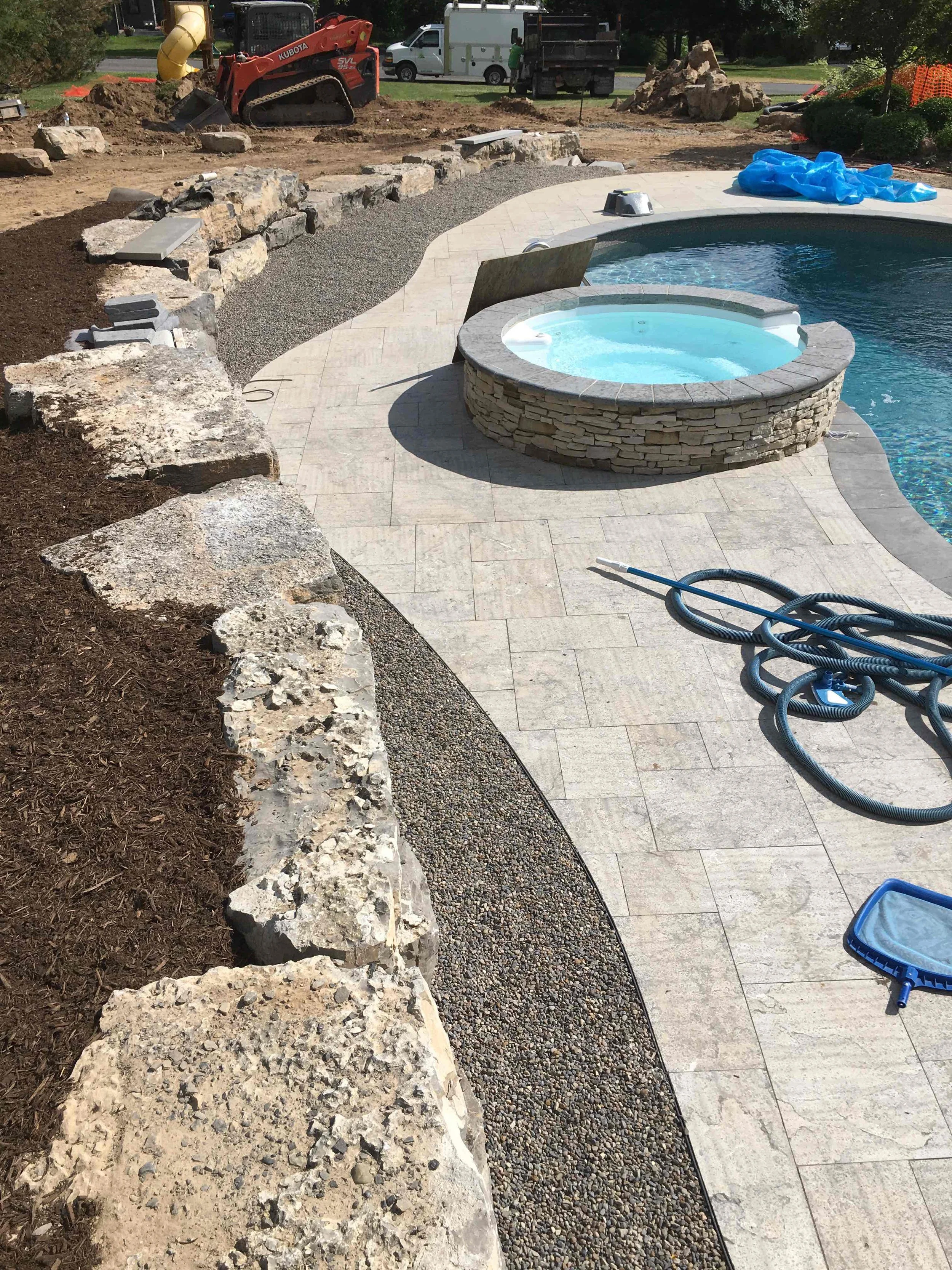 A backyard with a swimming pool and hot tub under construction. There are large rocks lining the edge, construction equipment, hoses, and some paving stones being installed.