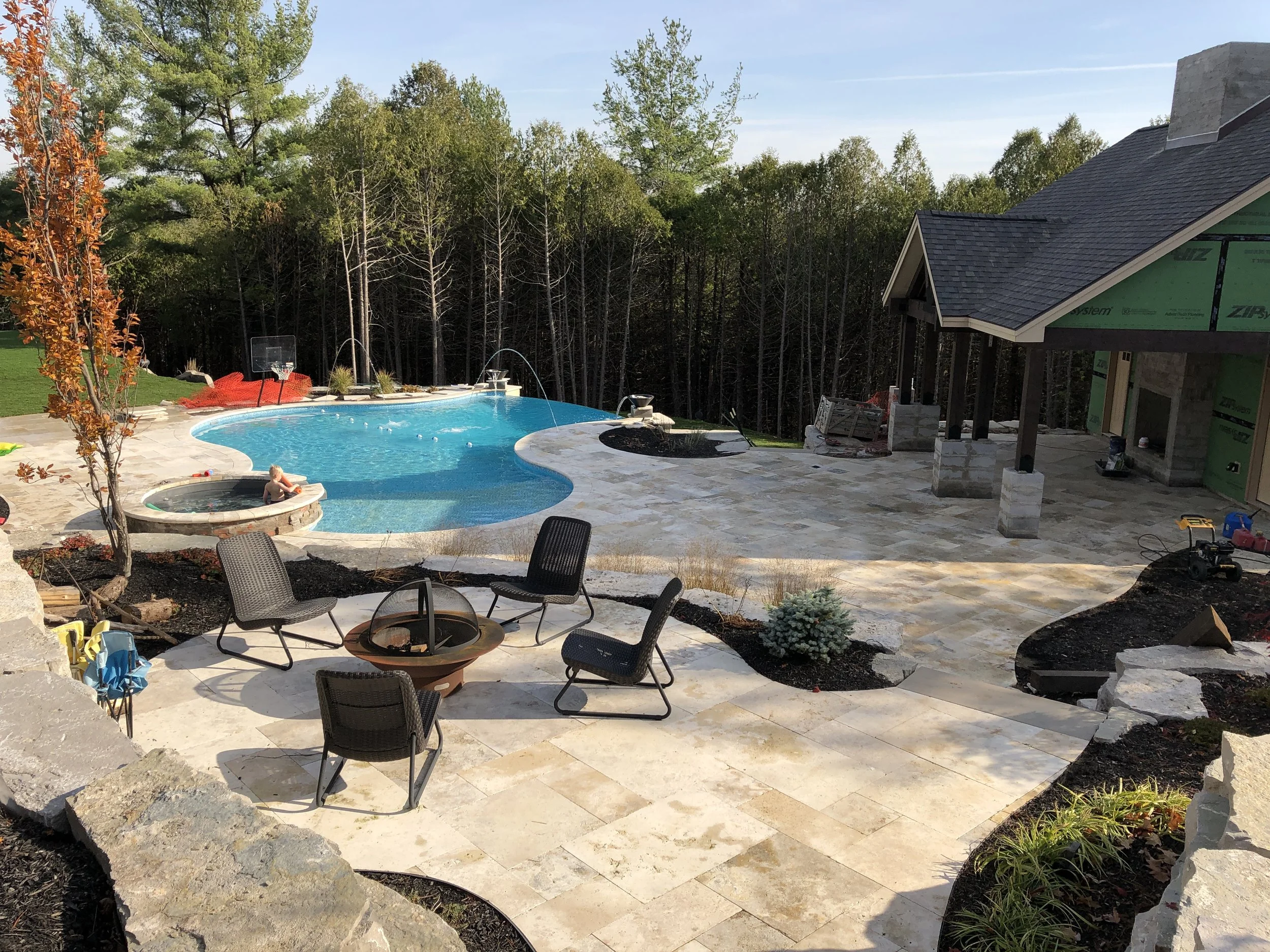 A backyard with a swimming pool, hot tub, and patio area under construction. There are four black chairs around a fire pit, and a small child is playing in the hot tub. In the background, a house is being built with visible green sheathing, and the a