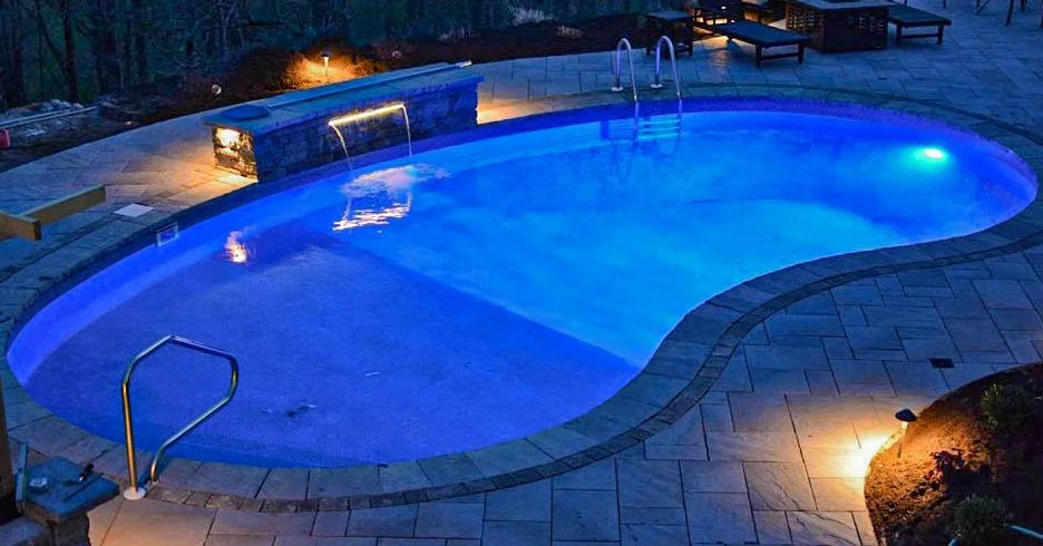 Nighttime view of an illuminated outdoor swimming pool with a curved shape, metal ladders, and surrounding poolside chairs and lighting.