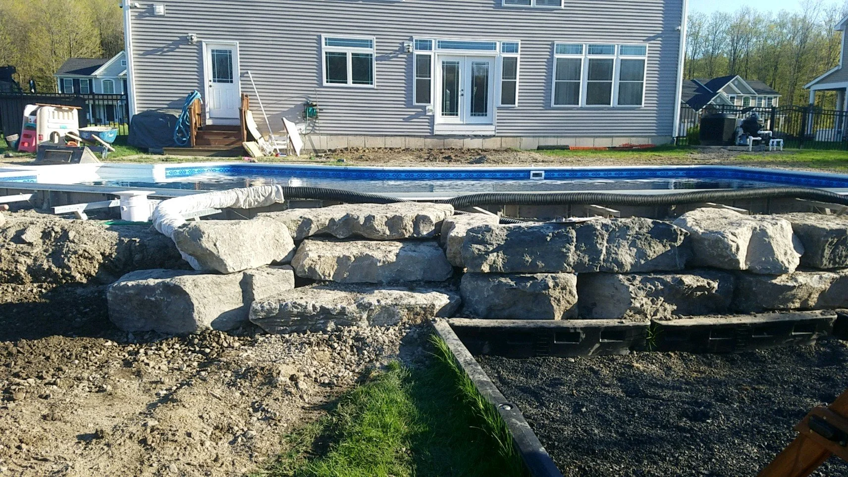 Backyard with pool under construction, stone wall being built in foreground, house with sliding glass door and multiple windows in background, outdoor furniture and toys on the patio.