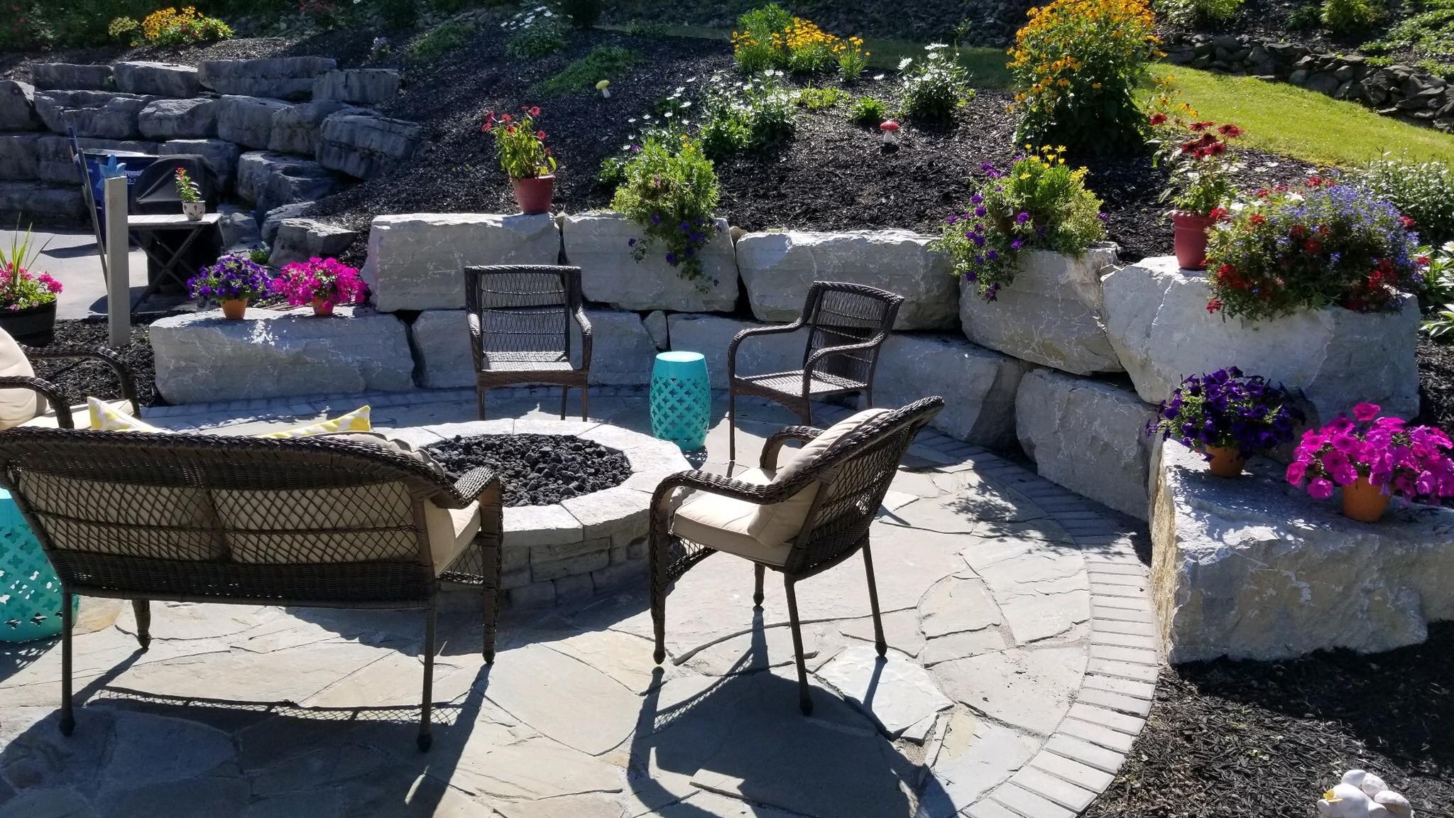 Outdoor patio with stone fire pit surrounded by chairs, potted flowers on large rocks, and a landscaped garden with various colorful flowers.