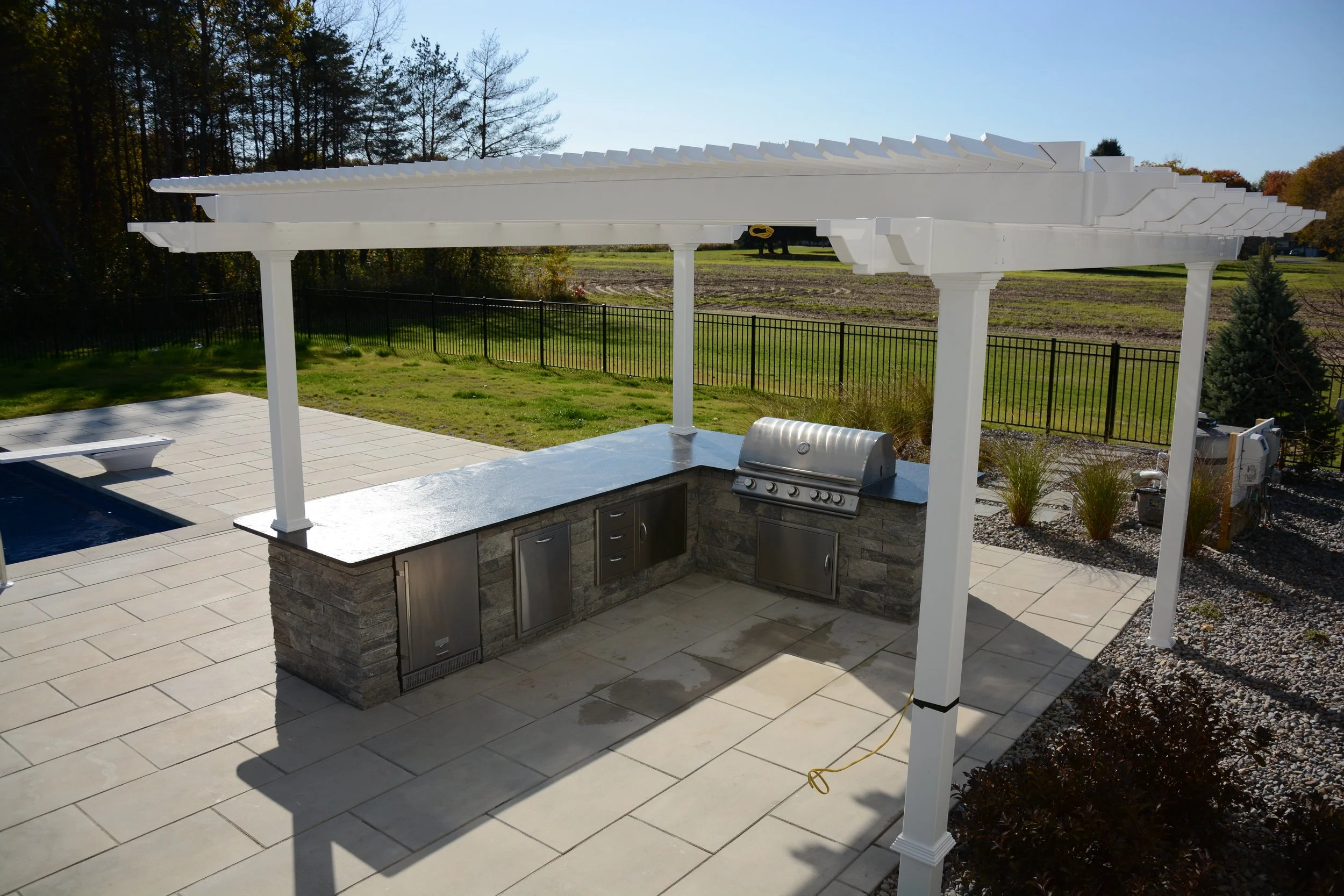Outdoor kitchen with a stone countertop, built-in grill, and storage cabinets, situated under a white pergola with lattice roof, overlooking a backyard with a pool, grassy area, trees, and a black metal fence.