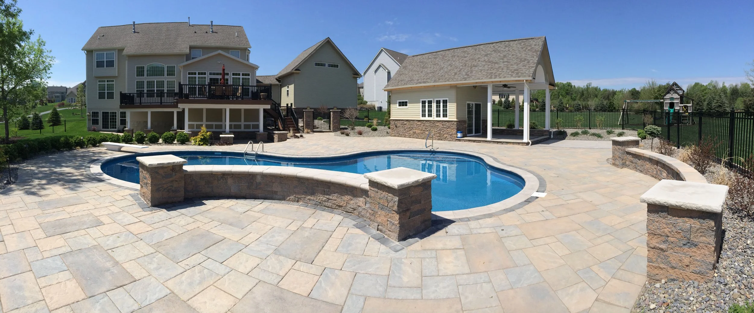 A backyard with a swimming pool, stone patio, a house with a deck, a pool house, and a child's play structure in a fenced yard.