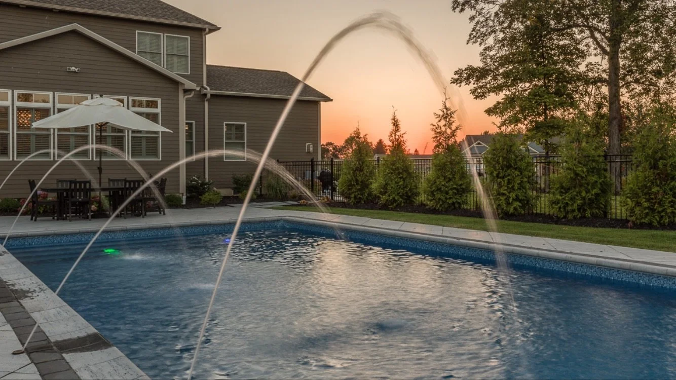A backyard swimming pool with water fountains during sunset, surrounded by a house, patio table with umbrella, trees, and a fence.