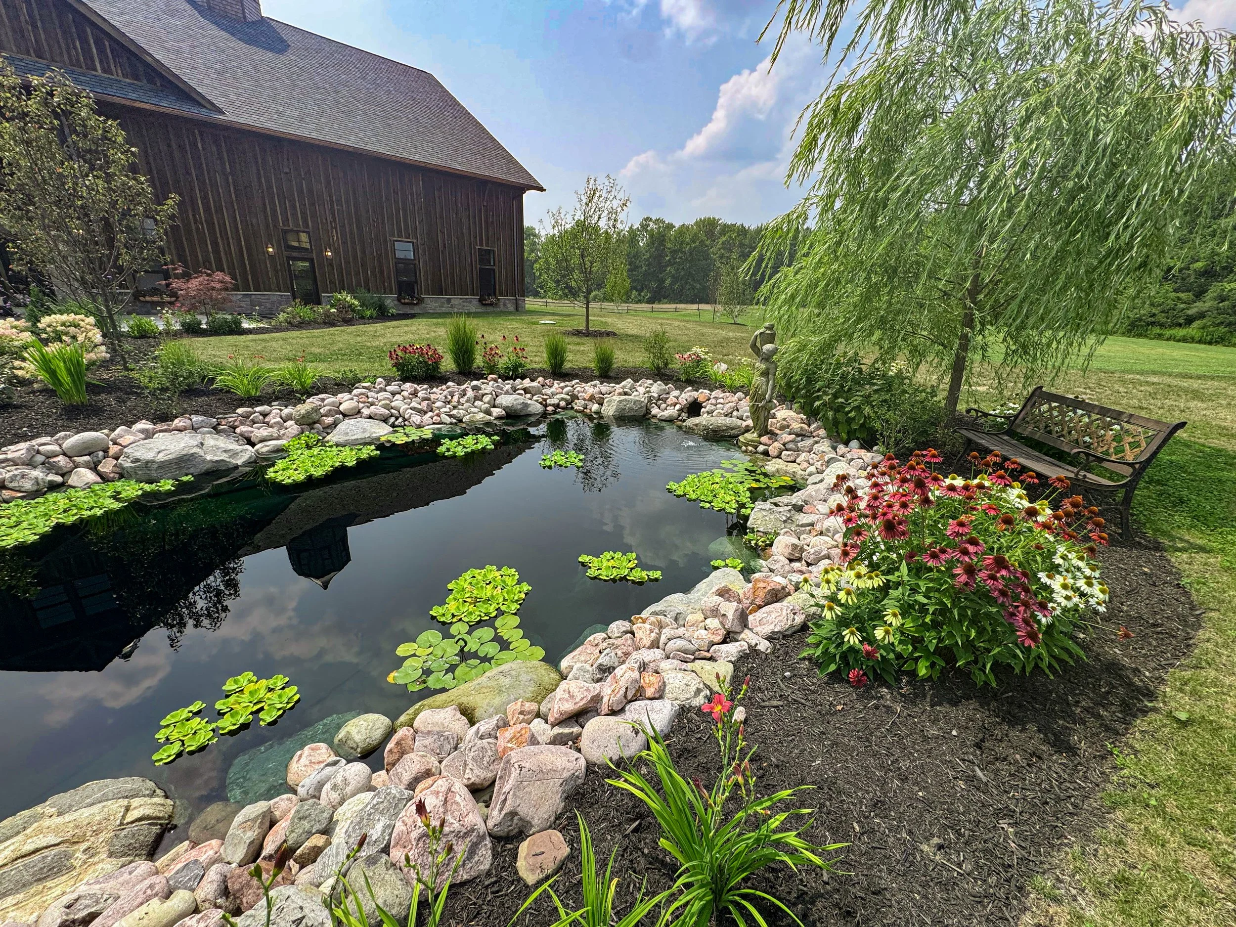 A backyard garden with a pond, surrounded by colorful flowers, rocks, and greenery, with a wooden barn in the background and a bench under a tree.