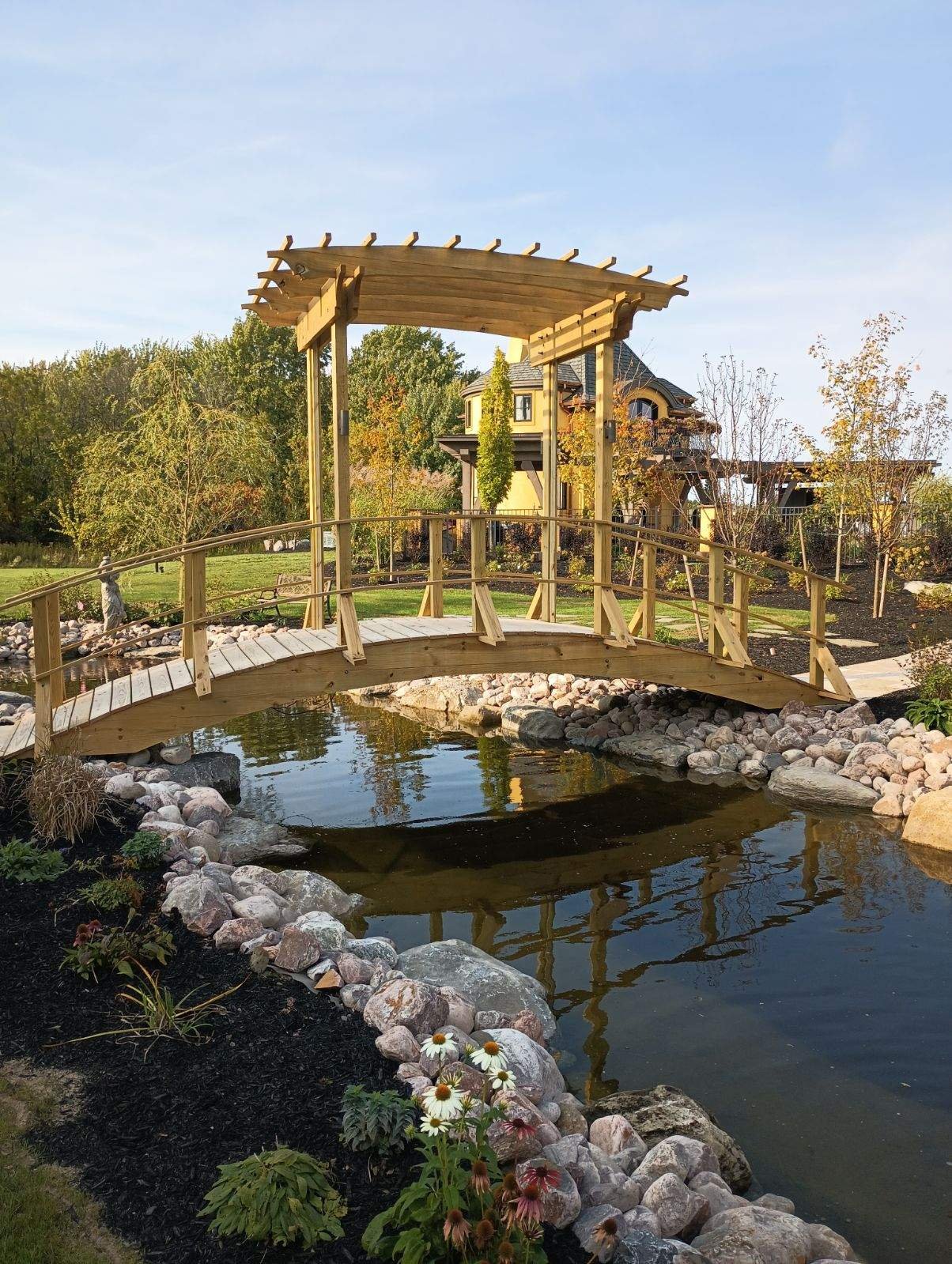 A wooden footbridge over a pond in a landscaped garden with trees, rocks, and a house in the background.
