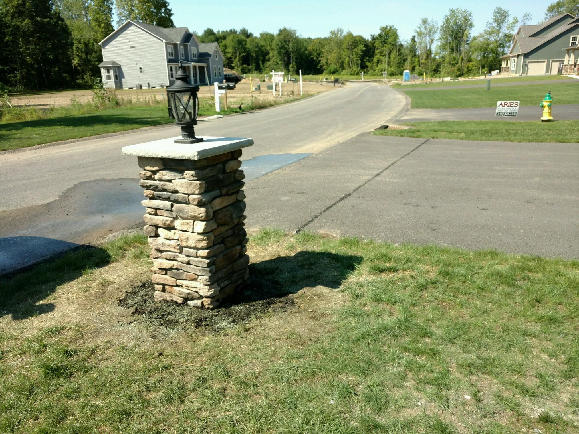 A stone and concrete pillar with a lantern on top near the edge of a grassy area, with a paved road, houses, and trees in the background.