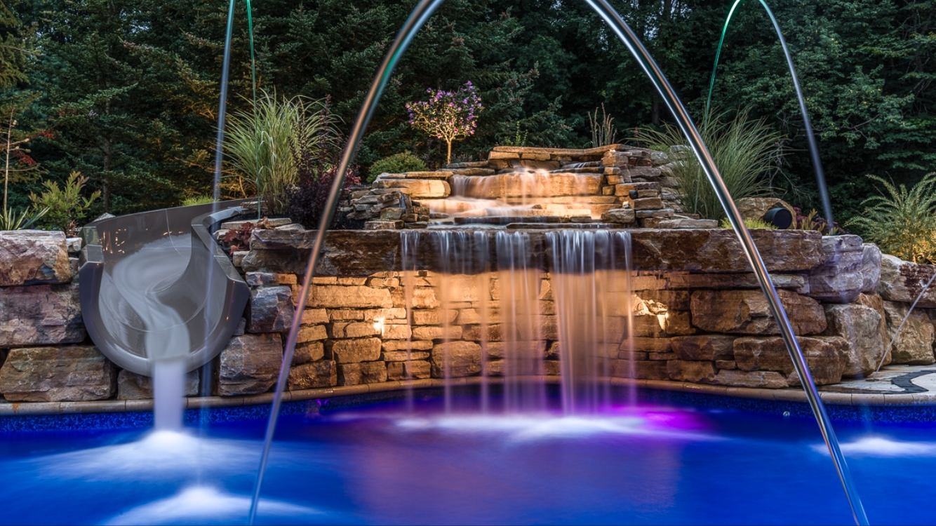 A backyard water feature with a waterfall cascading over stacked rocks into a pool, surrounded by lush plants and trees, with an illuminated slide and water jets.