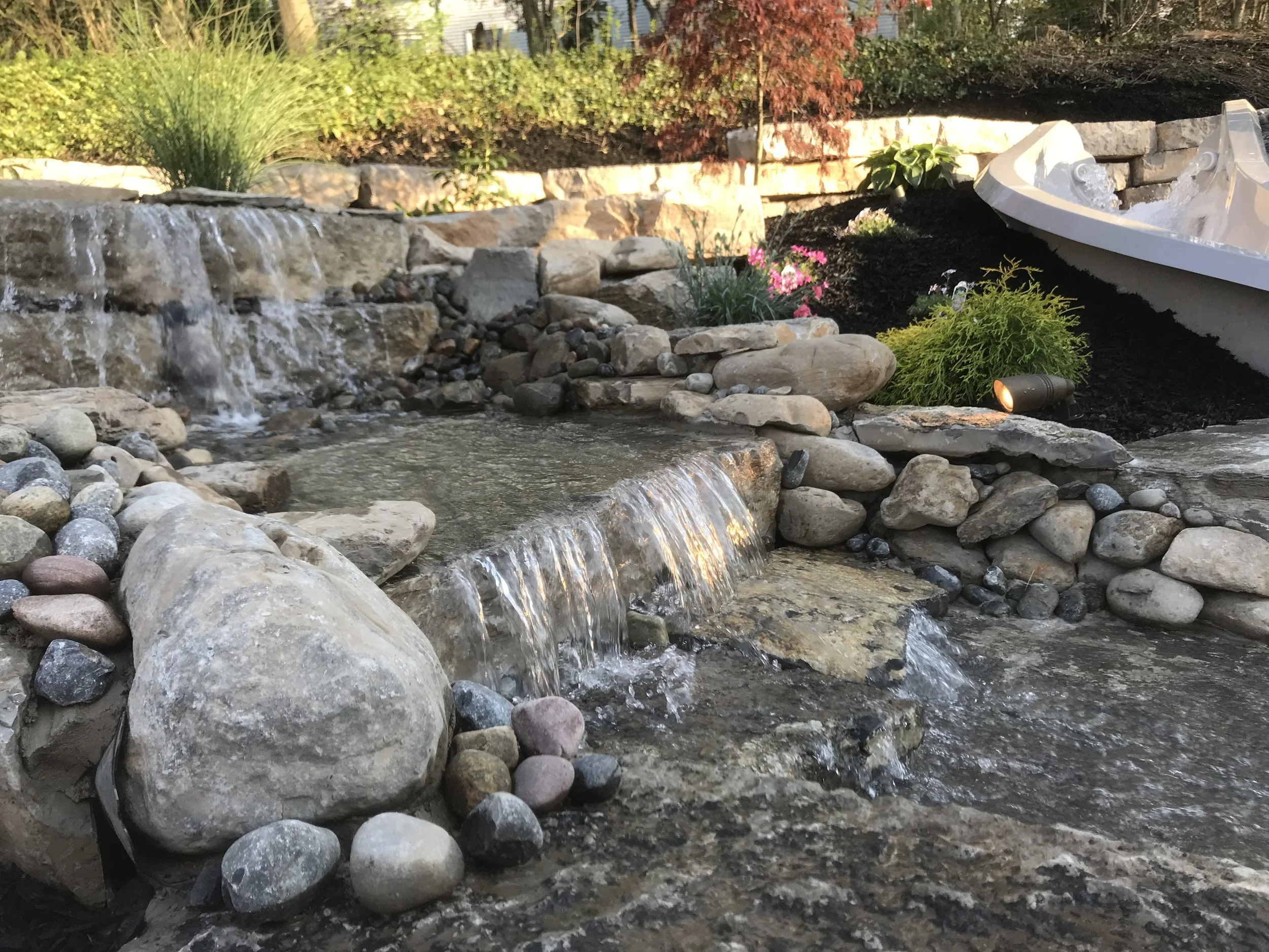 A small artificial waterfall with rocks and pebbles flowing into a pond, surrounded by garden plants and flowers.