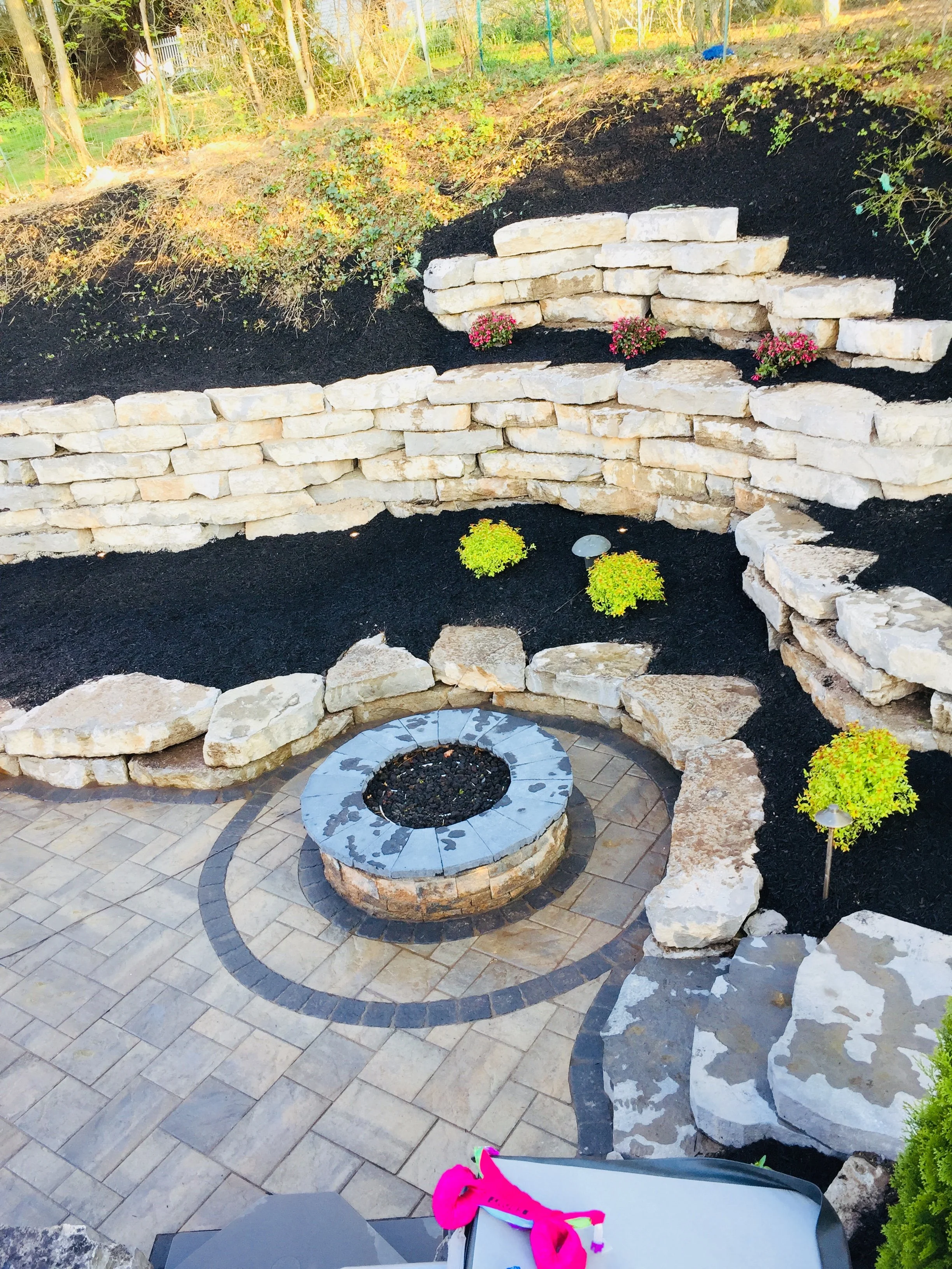 A landscaped backyard with a fire pit, stone retaining walls, and a patio with a circular design. Small plants and flowers are arranged around the area, with mulch and ladder-like stone steps leading up the hill.