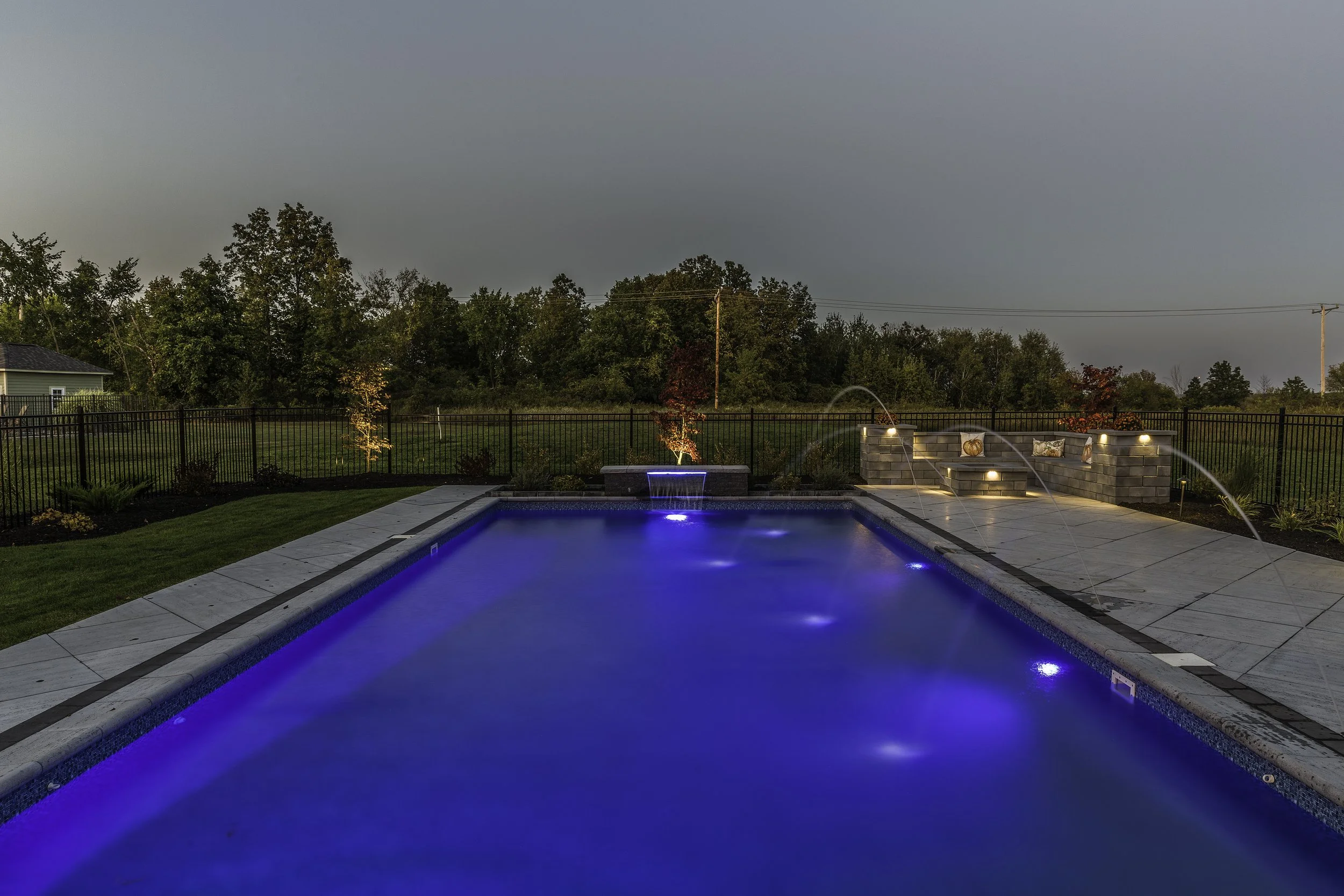 A backyard swimming pool at dusk illuminated with underwater and landscape lighting, surrounded by a deck and landscaped garden with trees and shrubs.
