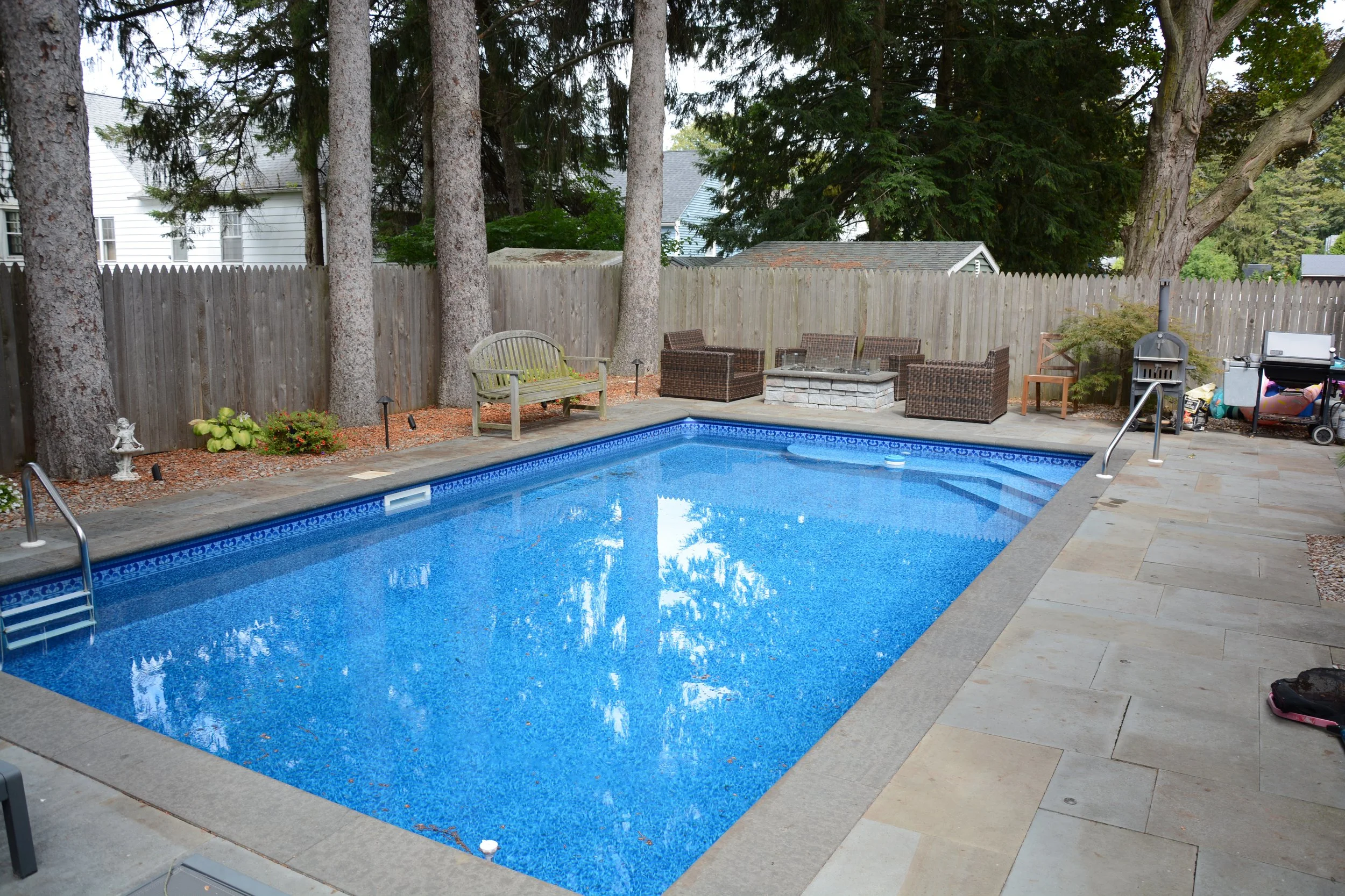 A backyard swimming pool with a stone deck, surrounded by a wooden fence and tall trees, with outdoor seating and a grill area.
