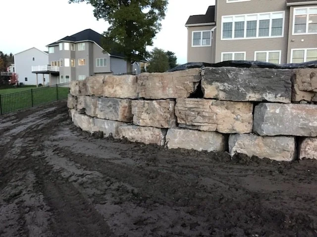 A dirt pathway beside a stone retaining wall in a residential area with multi-story apartment buildings in the background.