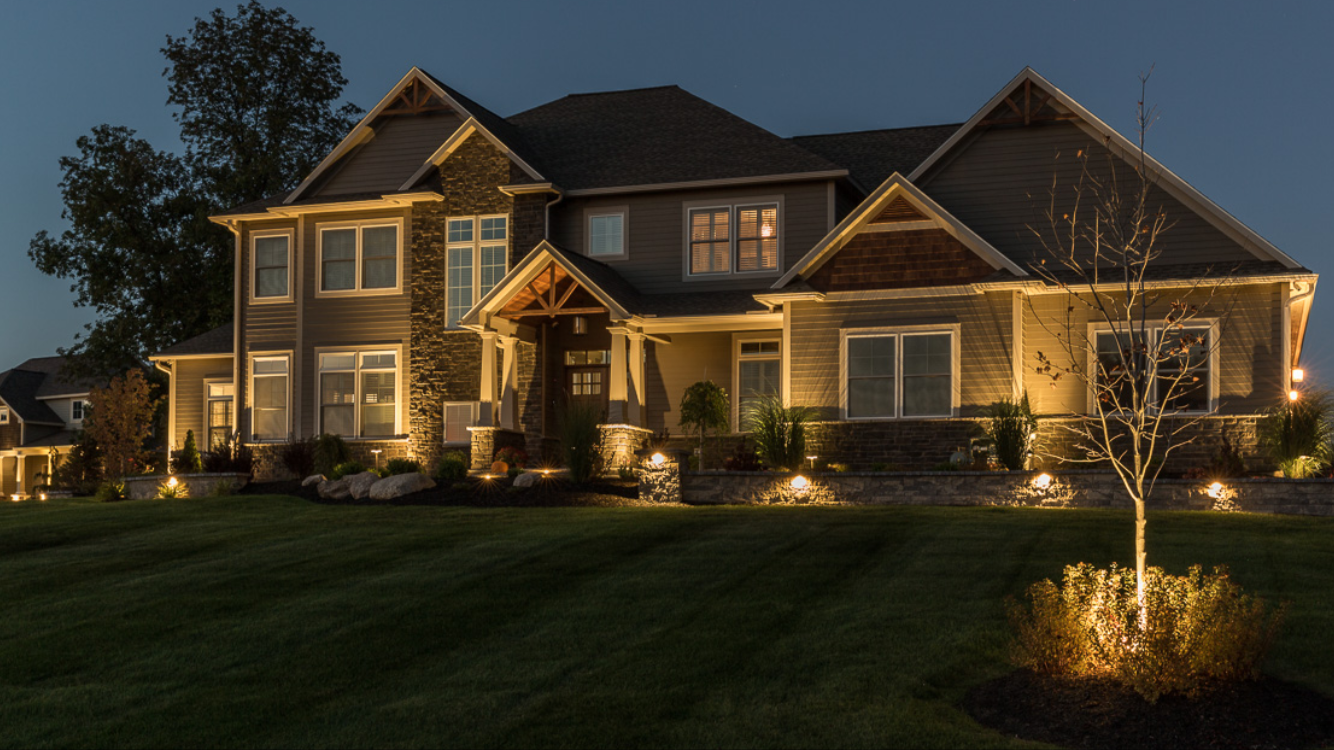 A large, modern house at night with exterior lighting illuminating the front yard, shrubs, and a tree.